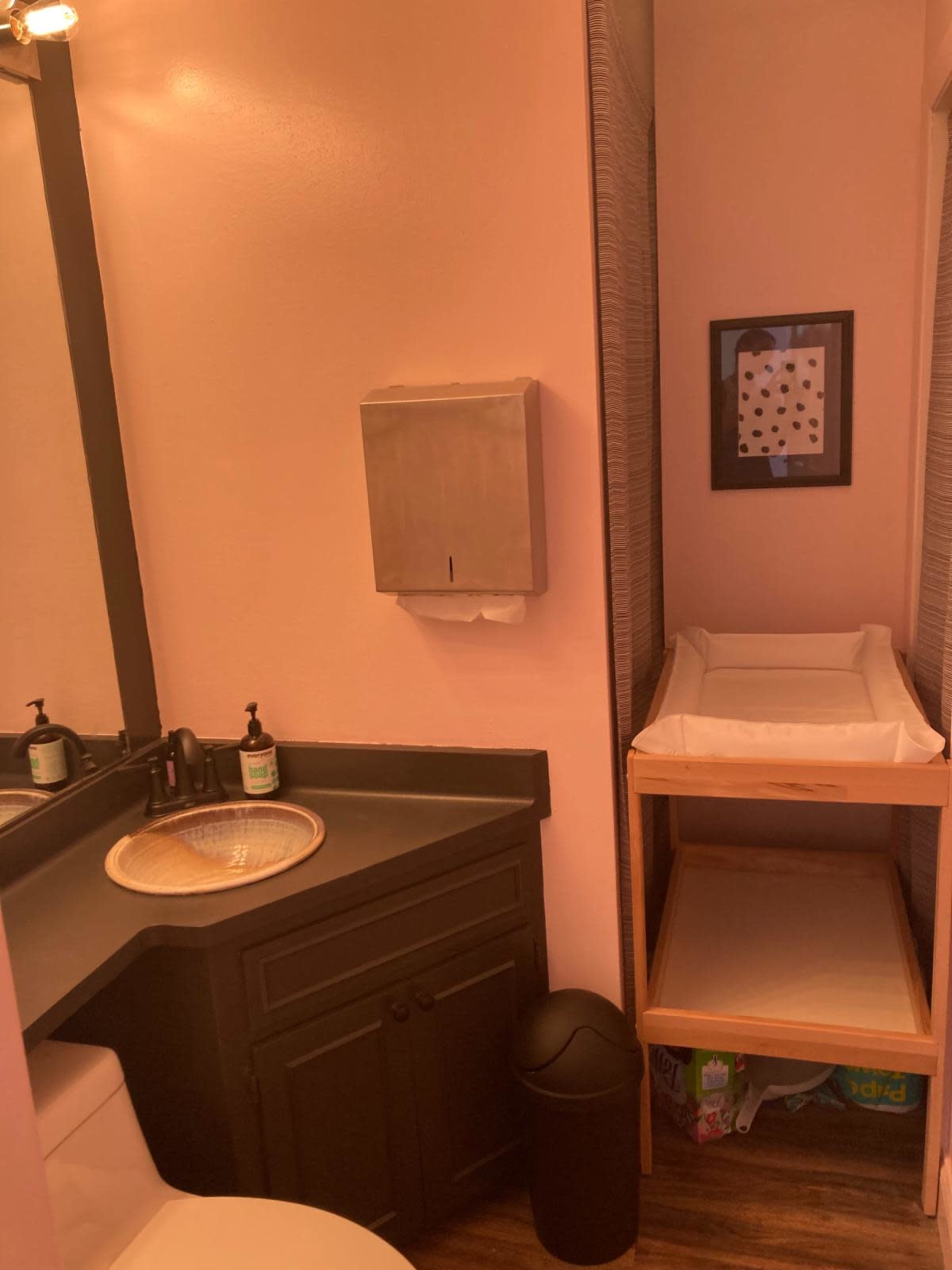 The image shows a small bathroom with a sink, a toilet, a changing table, and a mirror, all set against pink walls.
