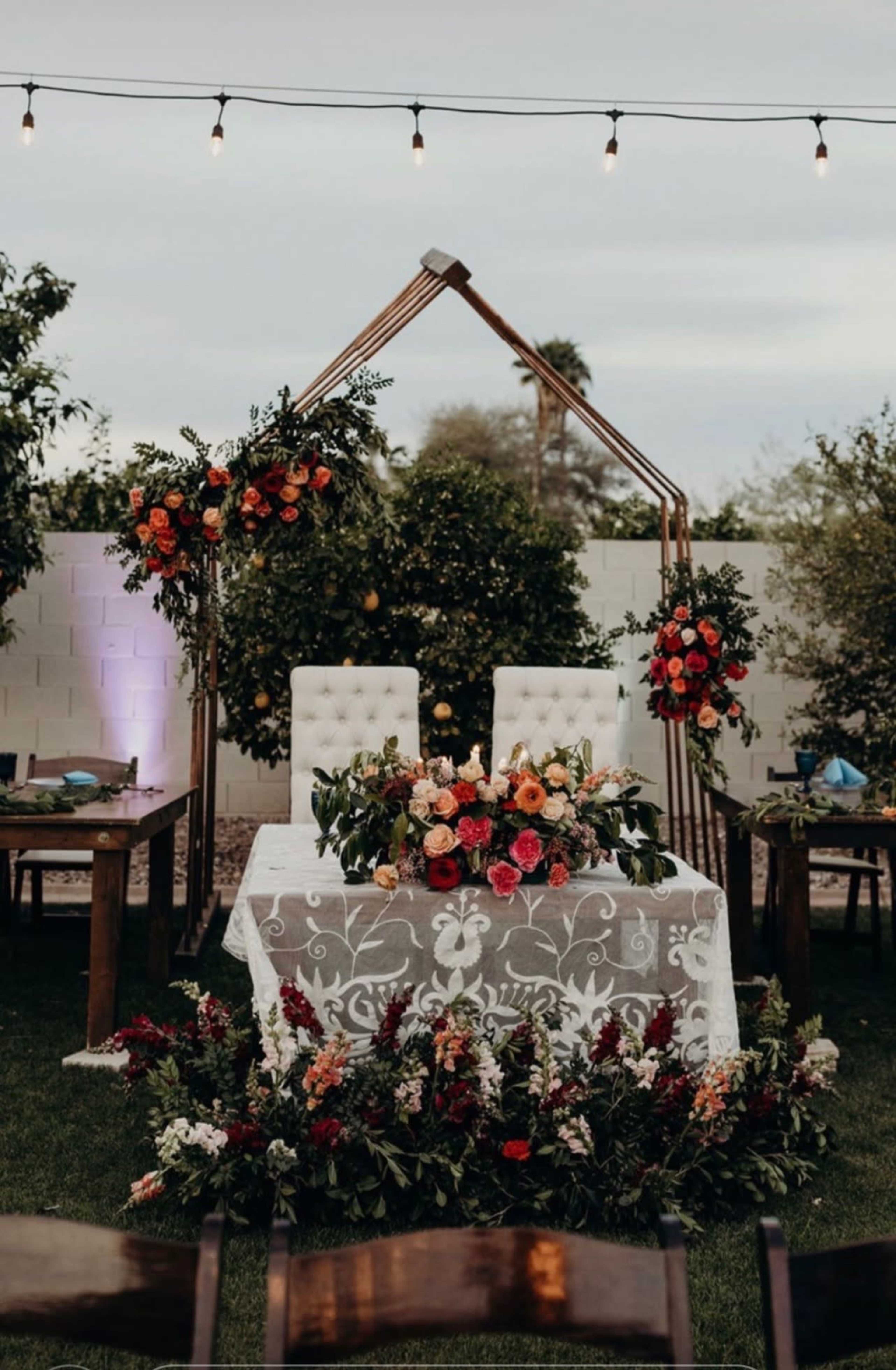 A decorated wedding altar features two elegant chairs flanked by floral arrangements and a white tablecloth adorned with a centerpiece of vibrant flowers.