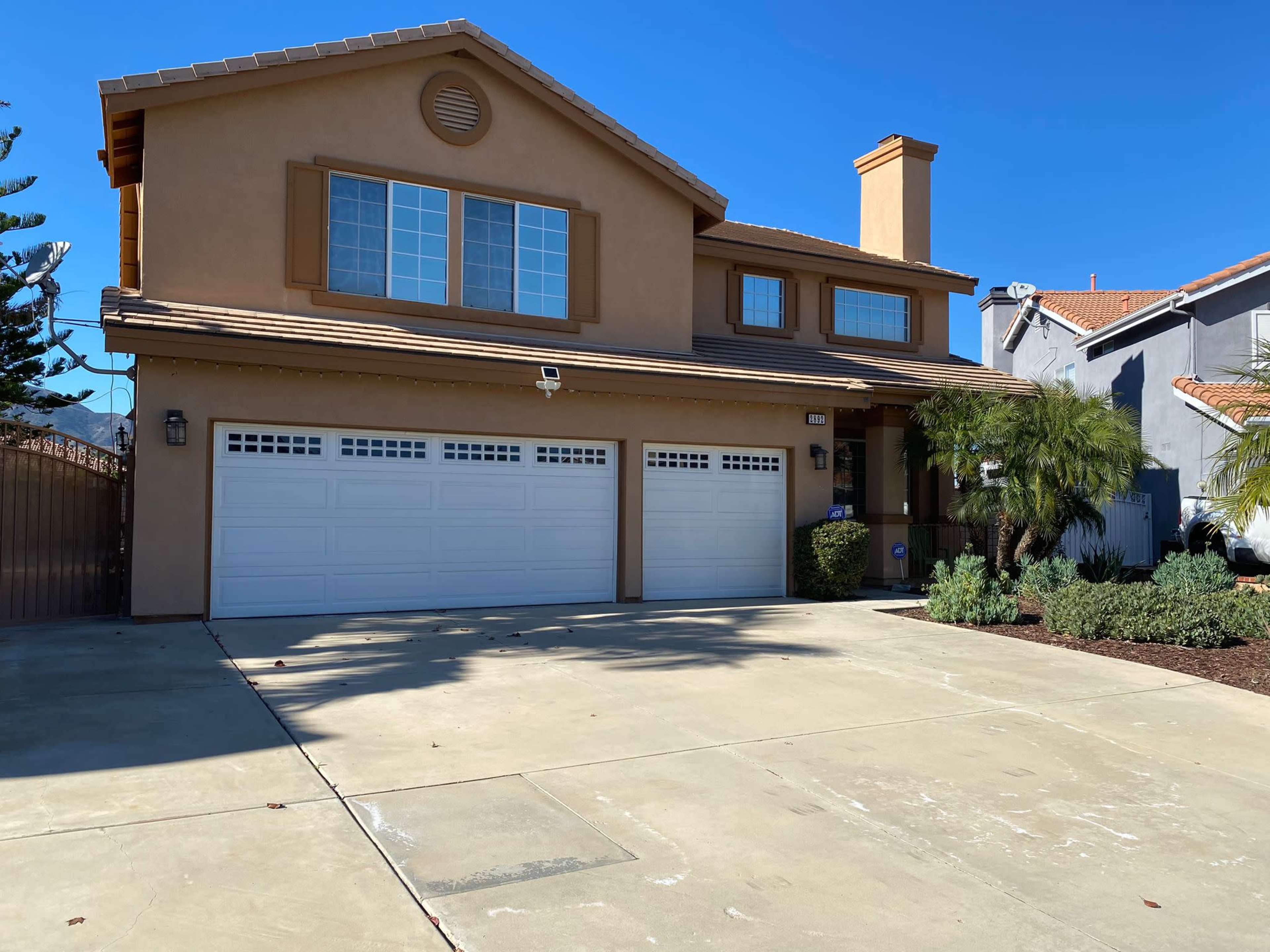The image shows a two-story residential house with a tan exterior, a sloped roof, and a three-car garage in front.