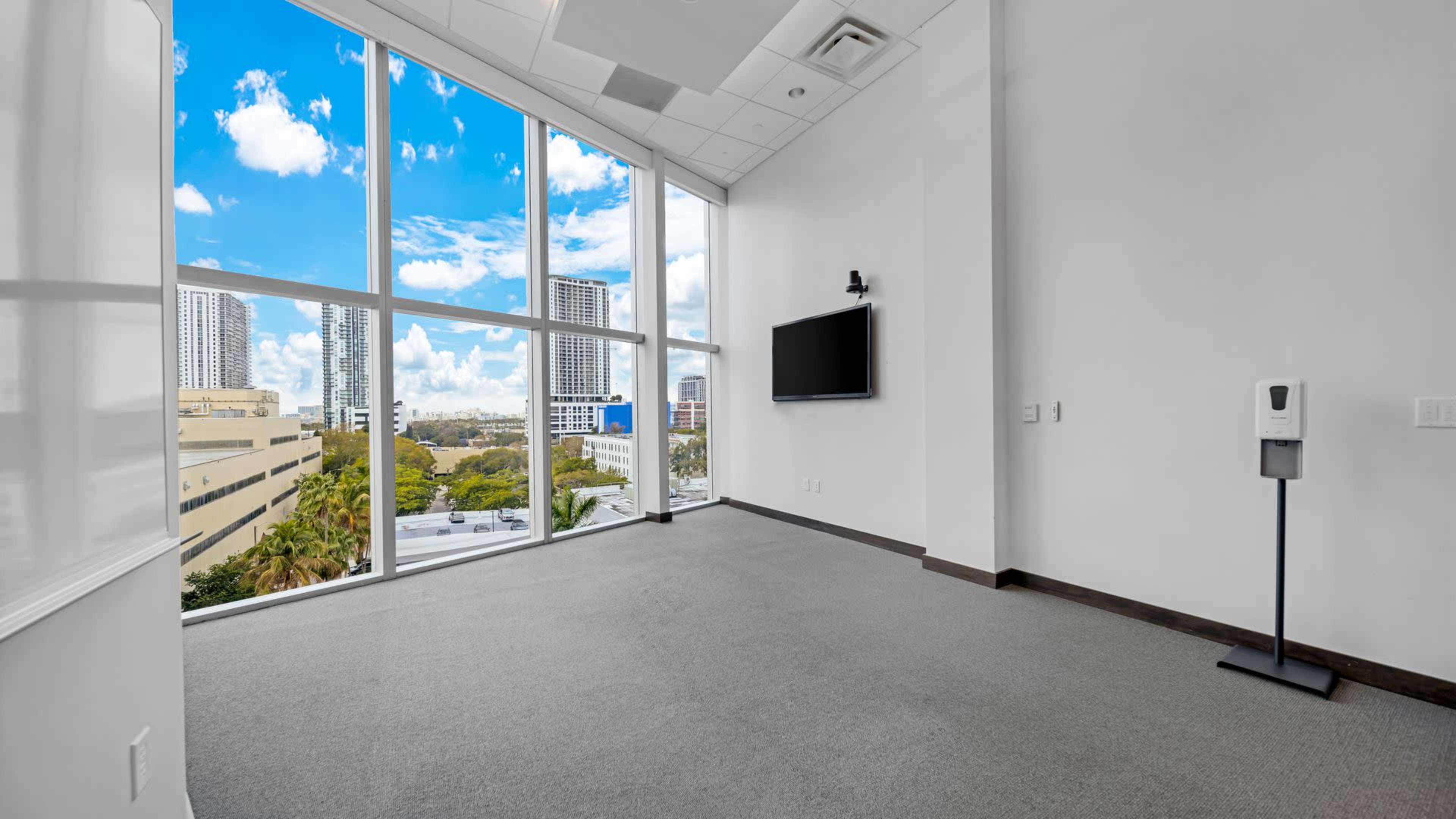 A spacious, empty room with large windows providing a view of a cityscape and a mounted television on the wall.