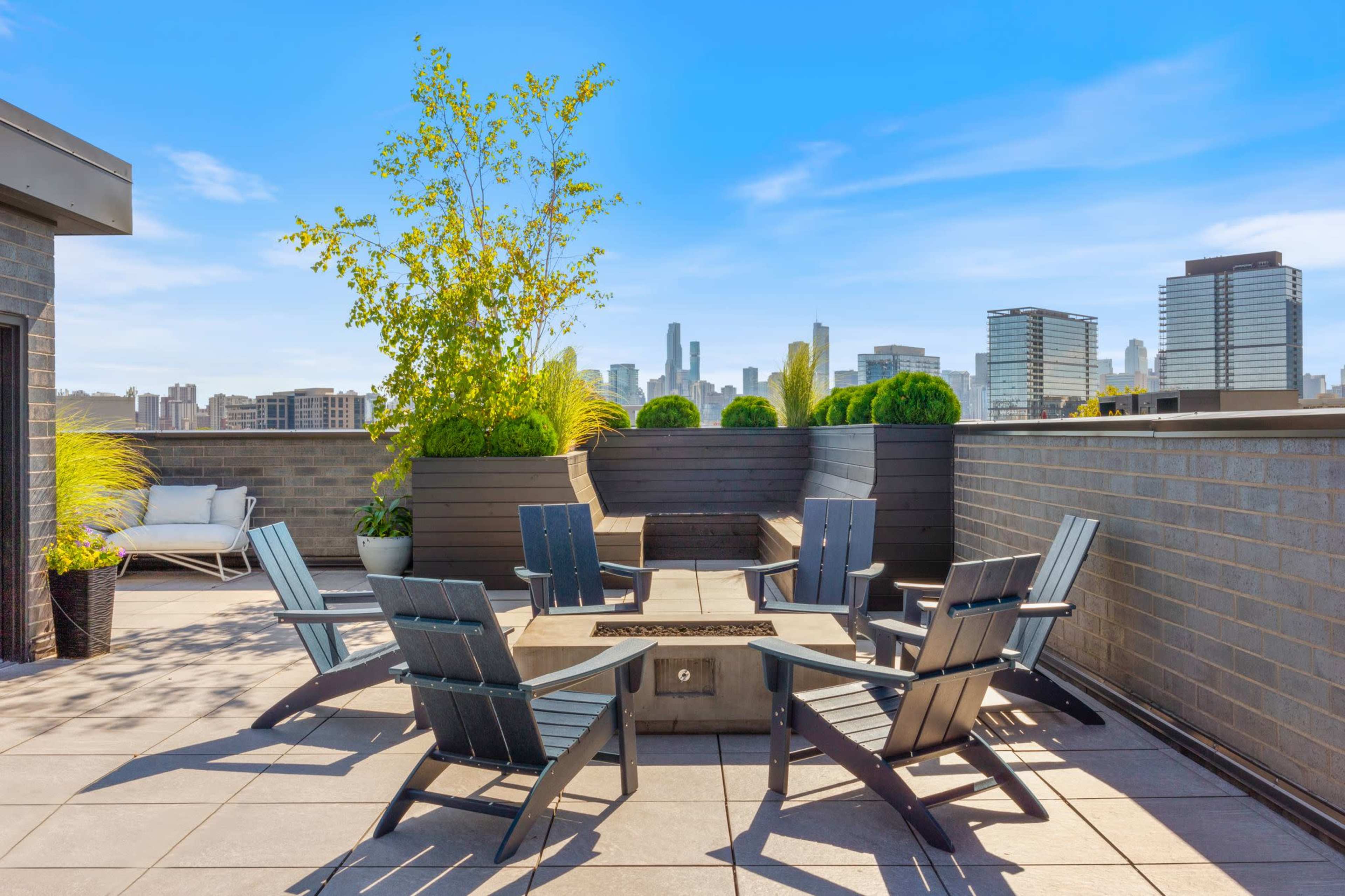 A rooftop terrace features a fire pit surrounded by several wooden chairs and planters with greenery, set against a skyline backdrop.
