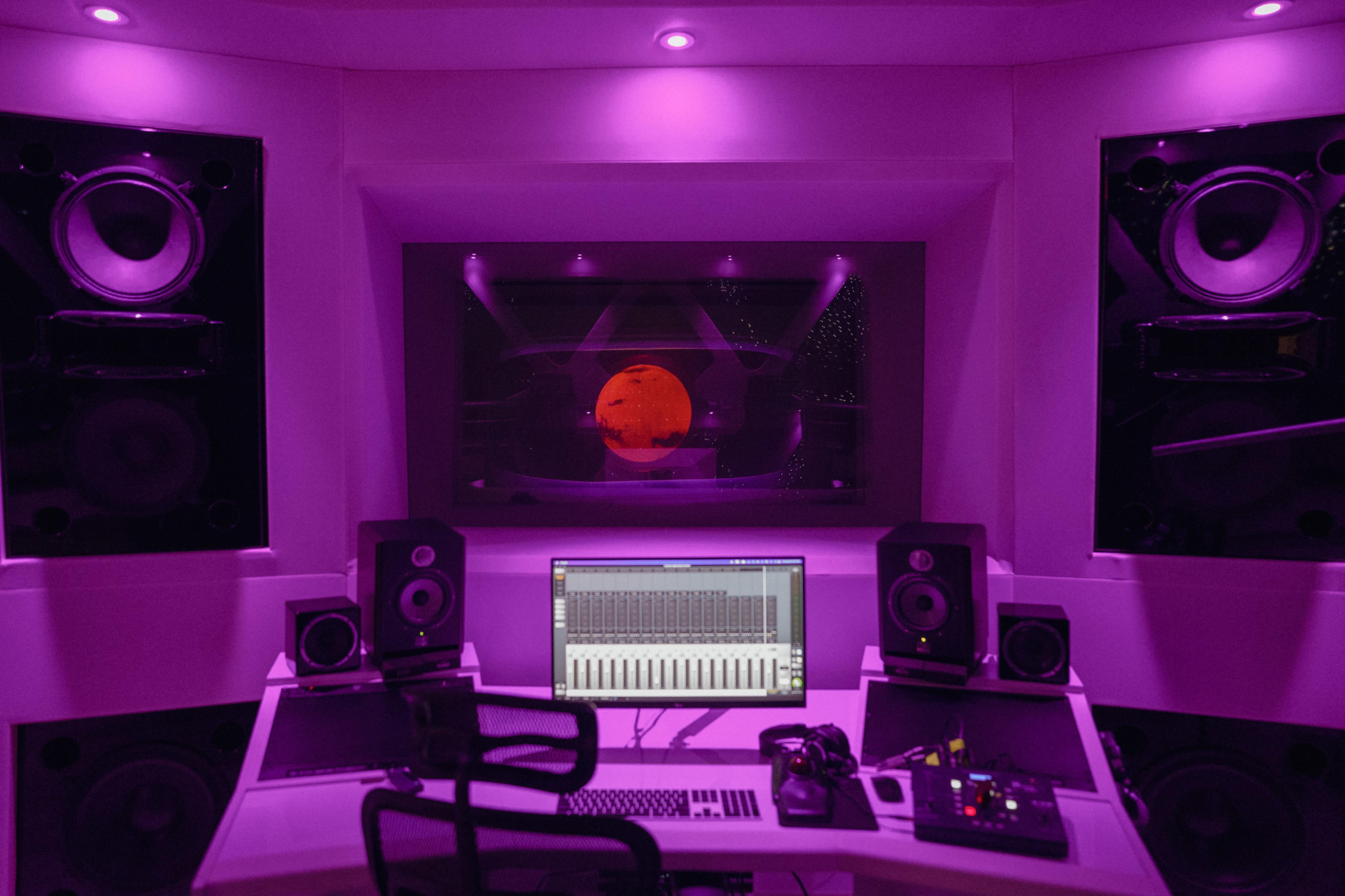 The image shows a modern music studio featuring a computer workstation, speakers, and a large window displaying a red planet against a starry background, all illuminated with purple lighting.