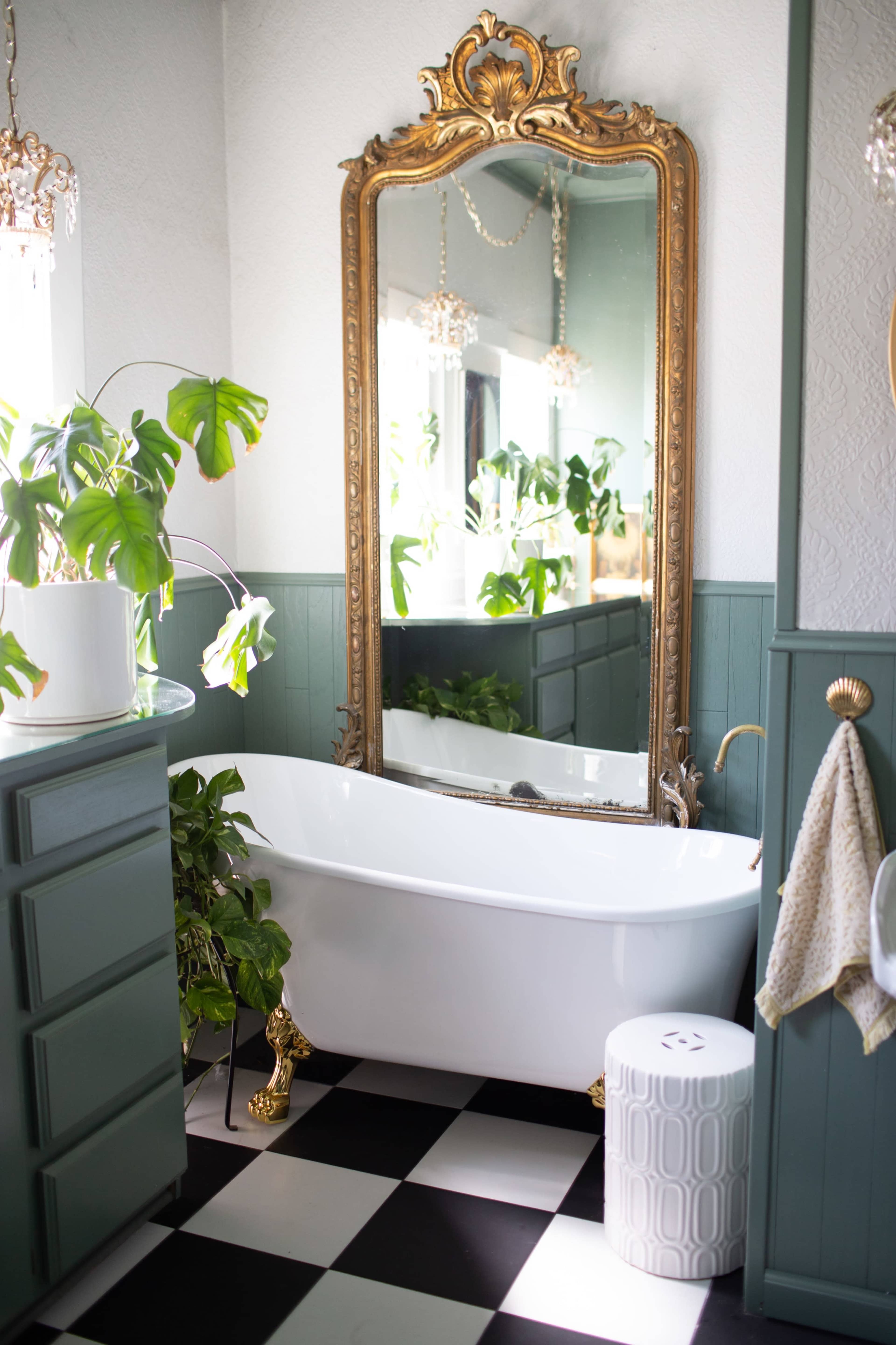 The image shows a bright bathroom with a freestanding white bathtub, a large ornate mirror, green plants, and a checkered black-and-white floor.