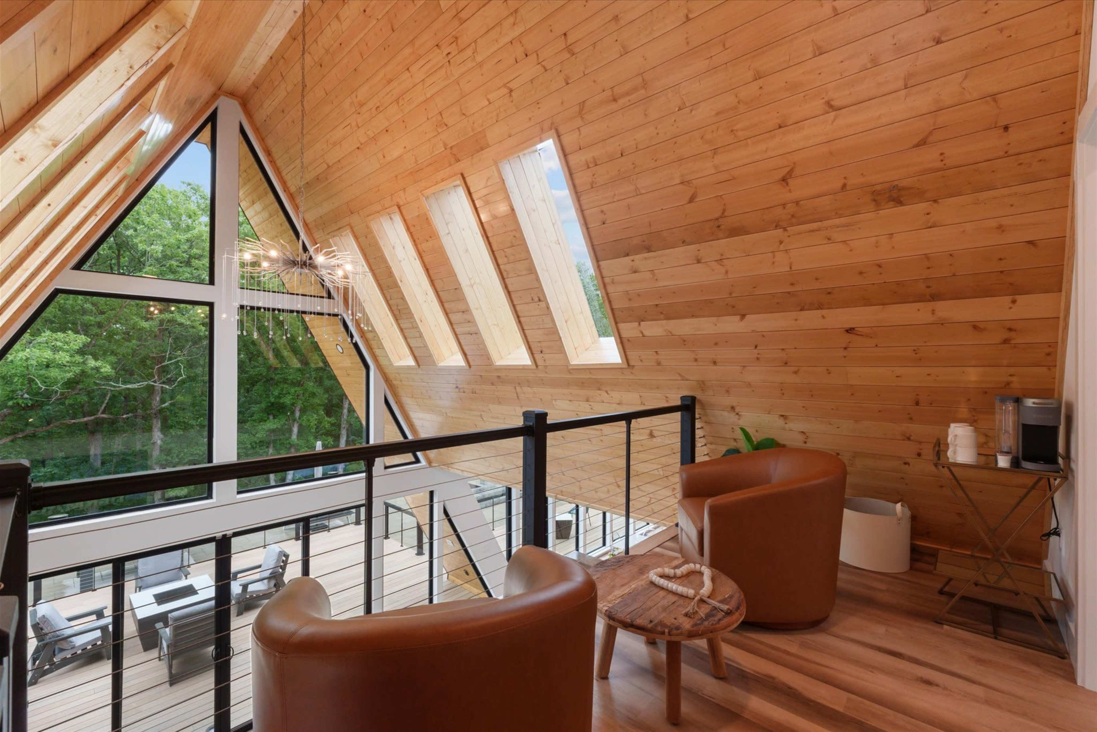 The image shows a modern wooden interior with an A-frame design, featuring two brown chairs and large windows overlooking greenery.