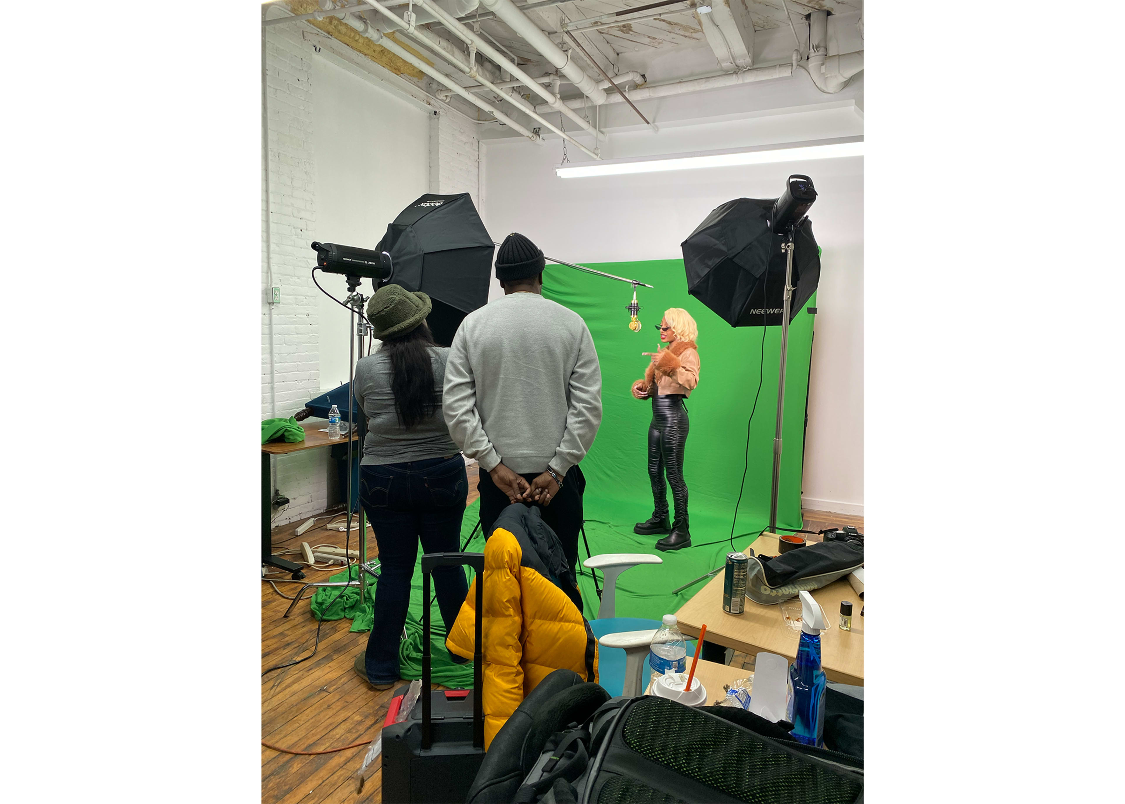 Three individuals are in a studio with a green screen, two at the back observing while one poses in front of the cameras with lighting equipment set up.