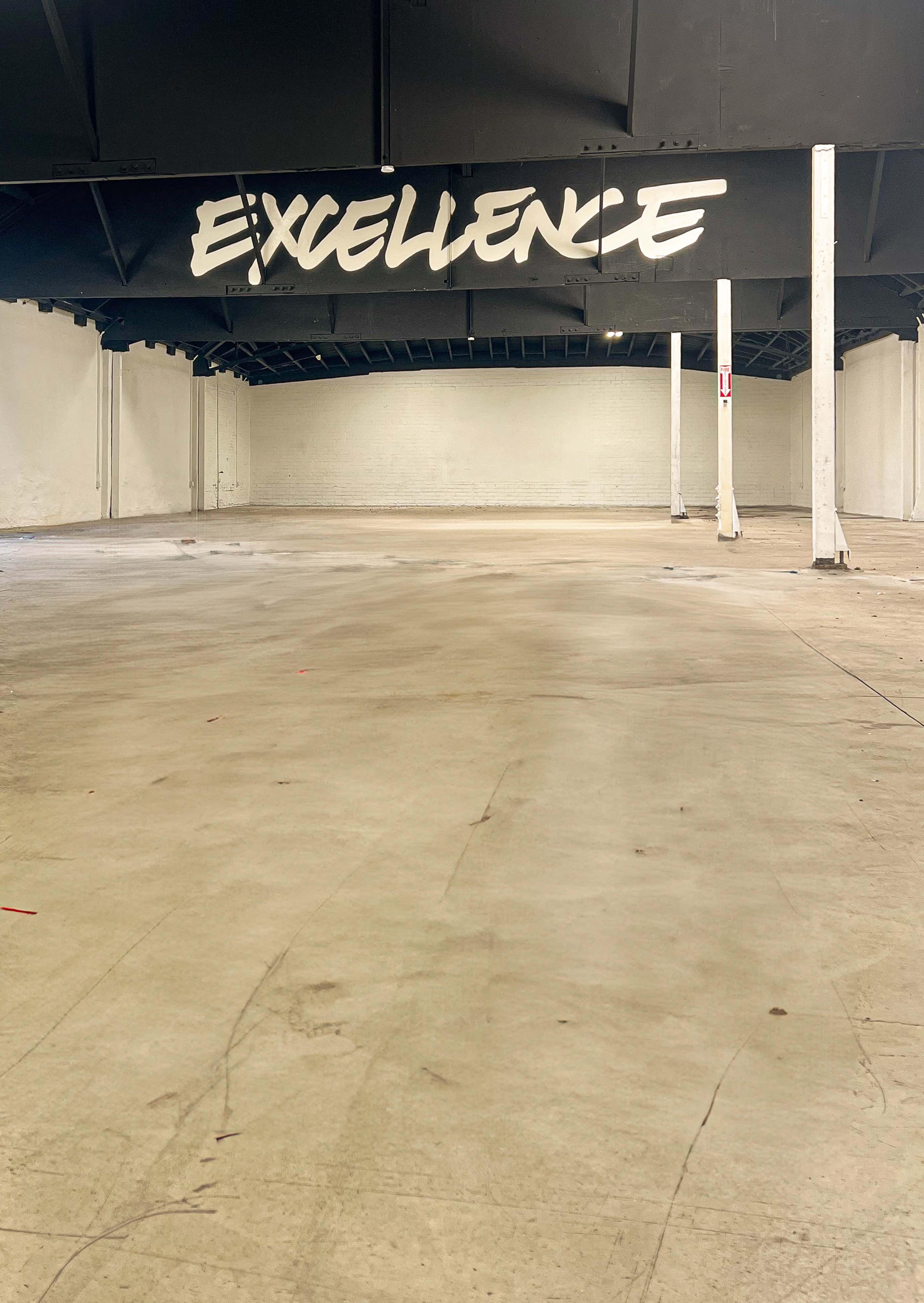 The image shows a spacious, empty warehouse with the word "EXCELLENCE" painted on the ceiling.