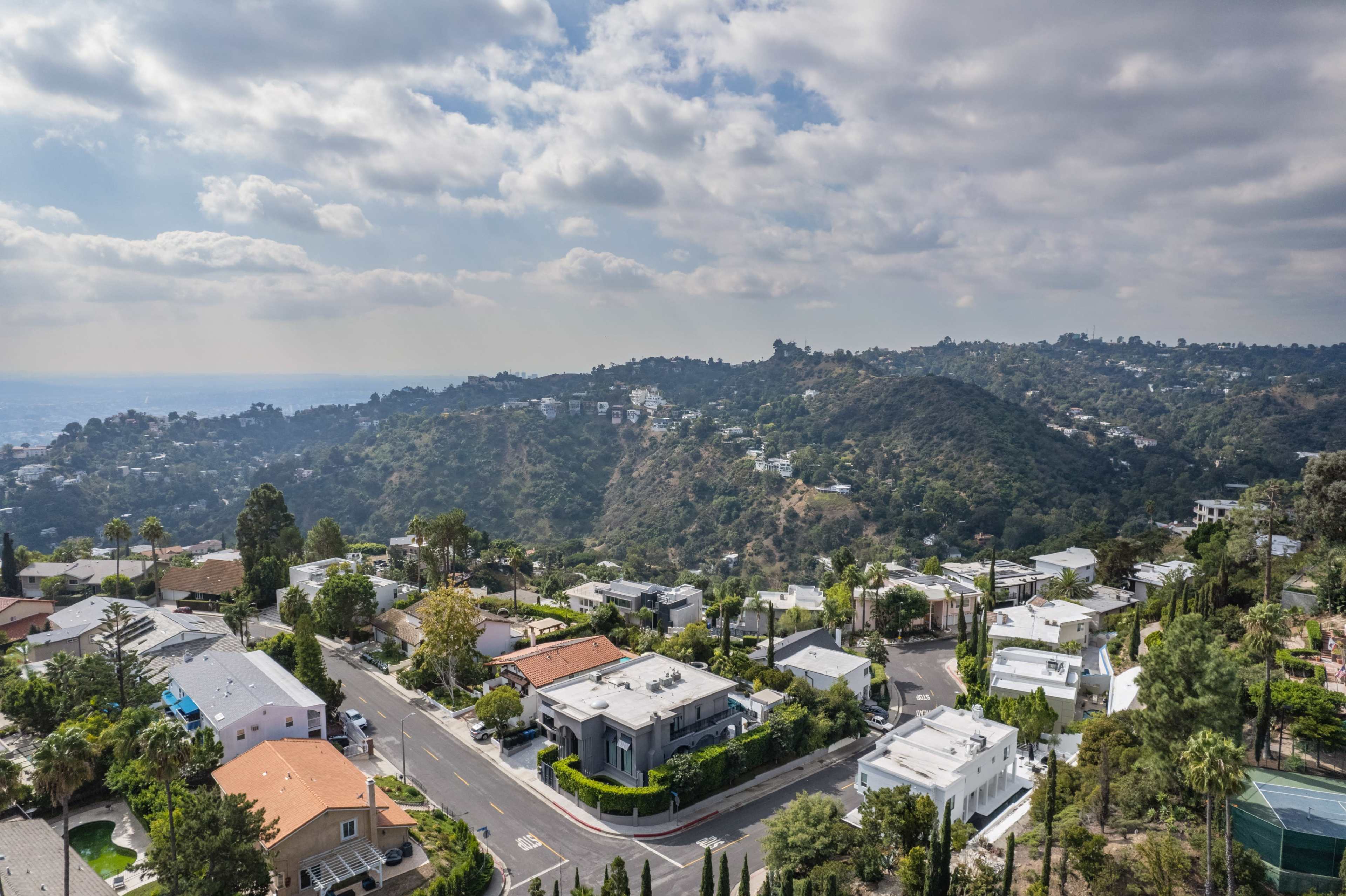 The image shows aerial views of residential homes and hilly terrain in a suburban area, under a partly cloudy sky.