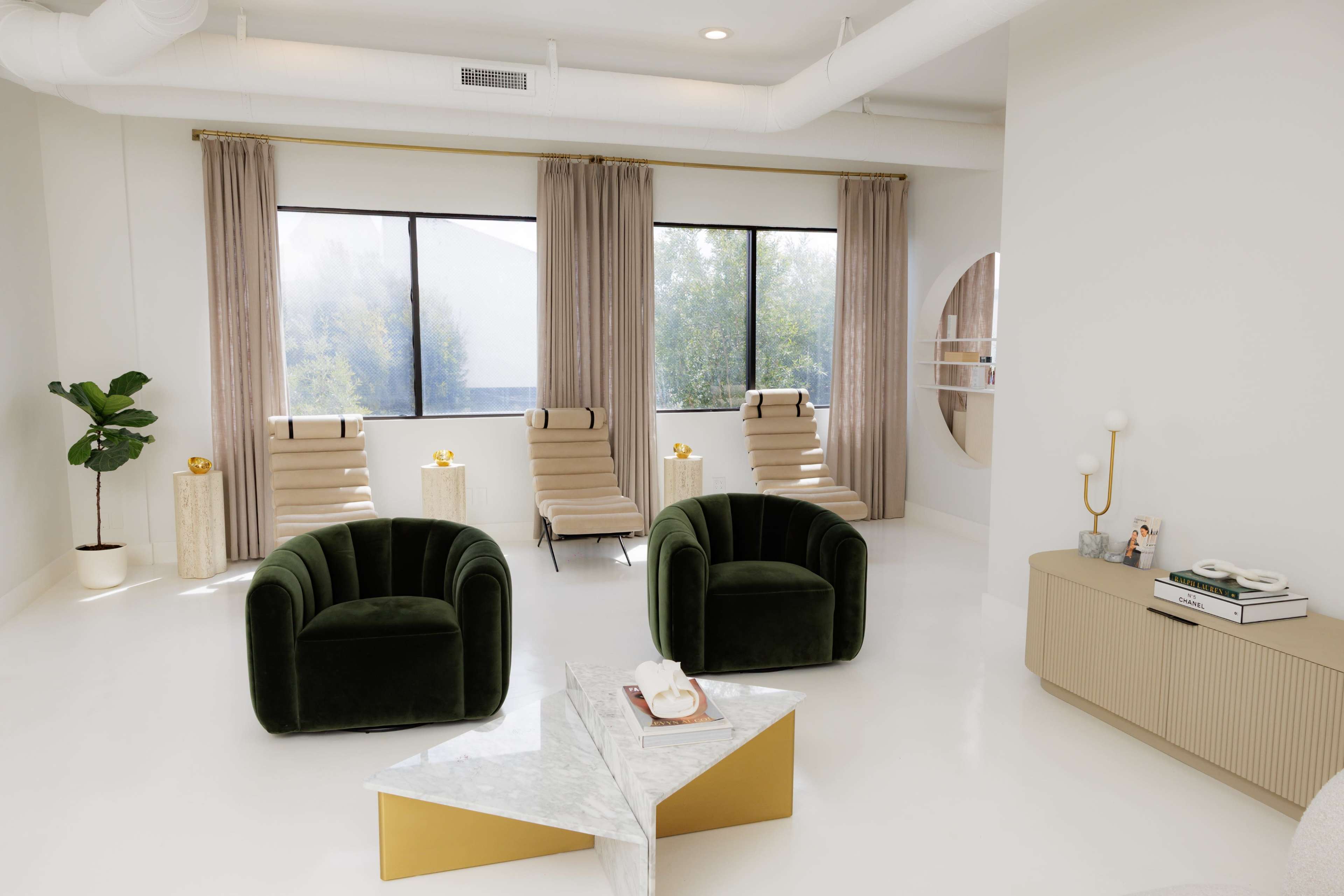 A bright and modern lounge area features two green velvet chairs, light-colored walls, and large windows with sheer curtains allowing natural light to enter.