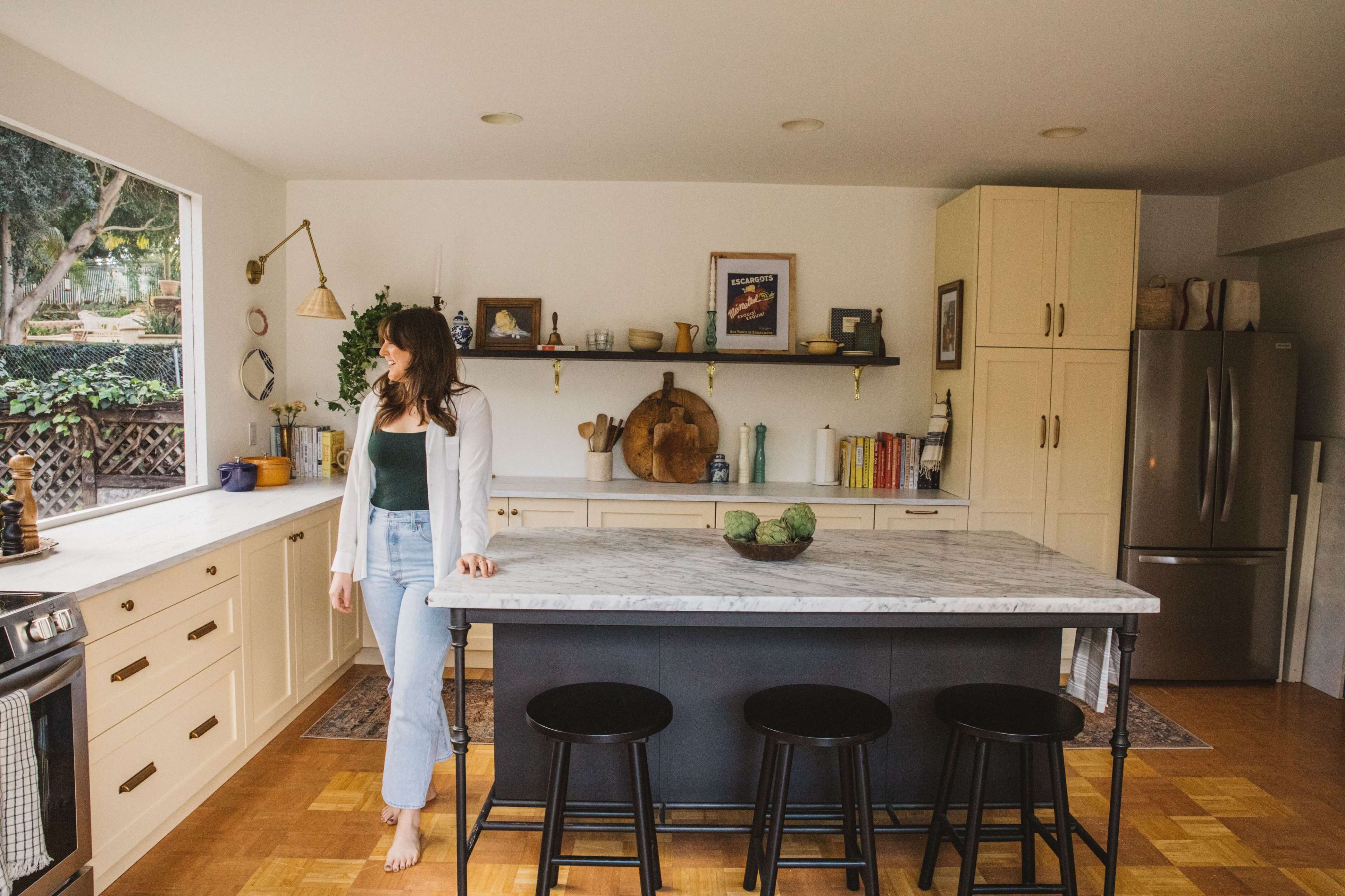 A woman stands near a marble-topped kitchen island in a bright kitchen with wooden floors and minimalist decor.