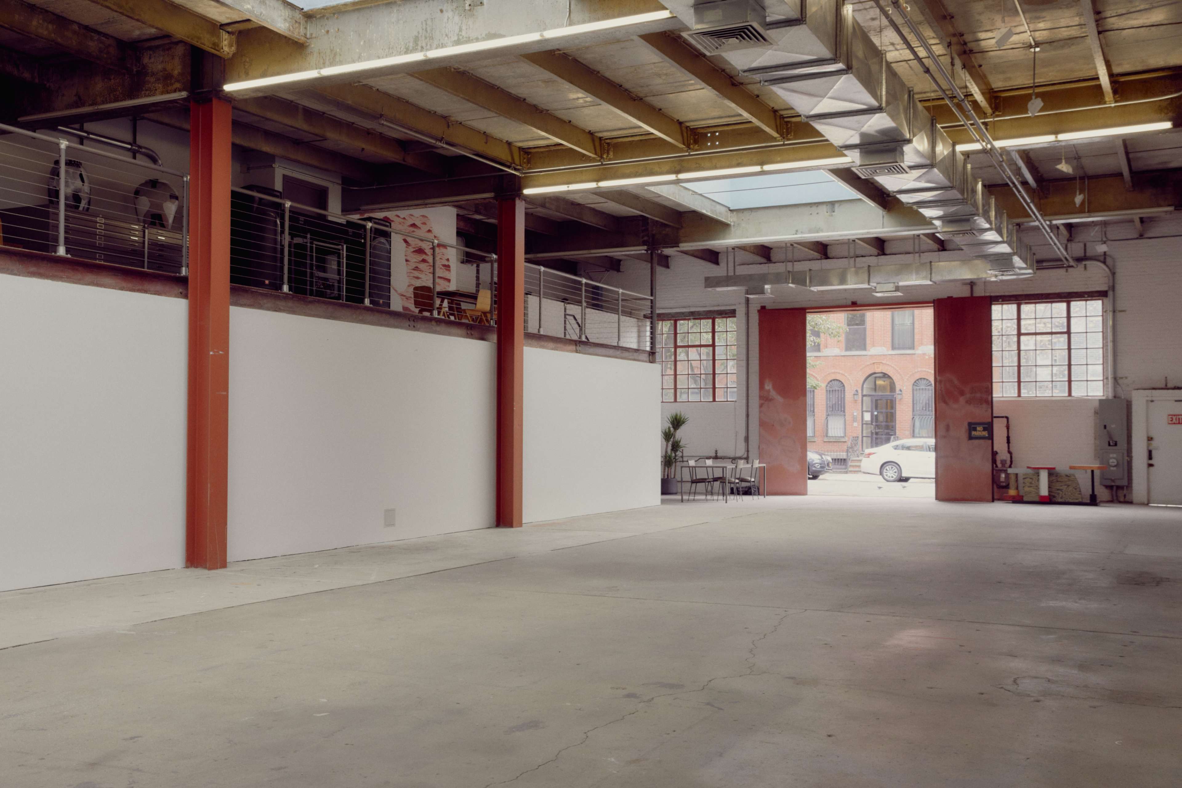 The image shows an empty industrial space with exposed beams and large windows, leading to an exterior view of a street and buildings.