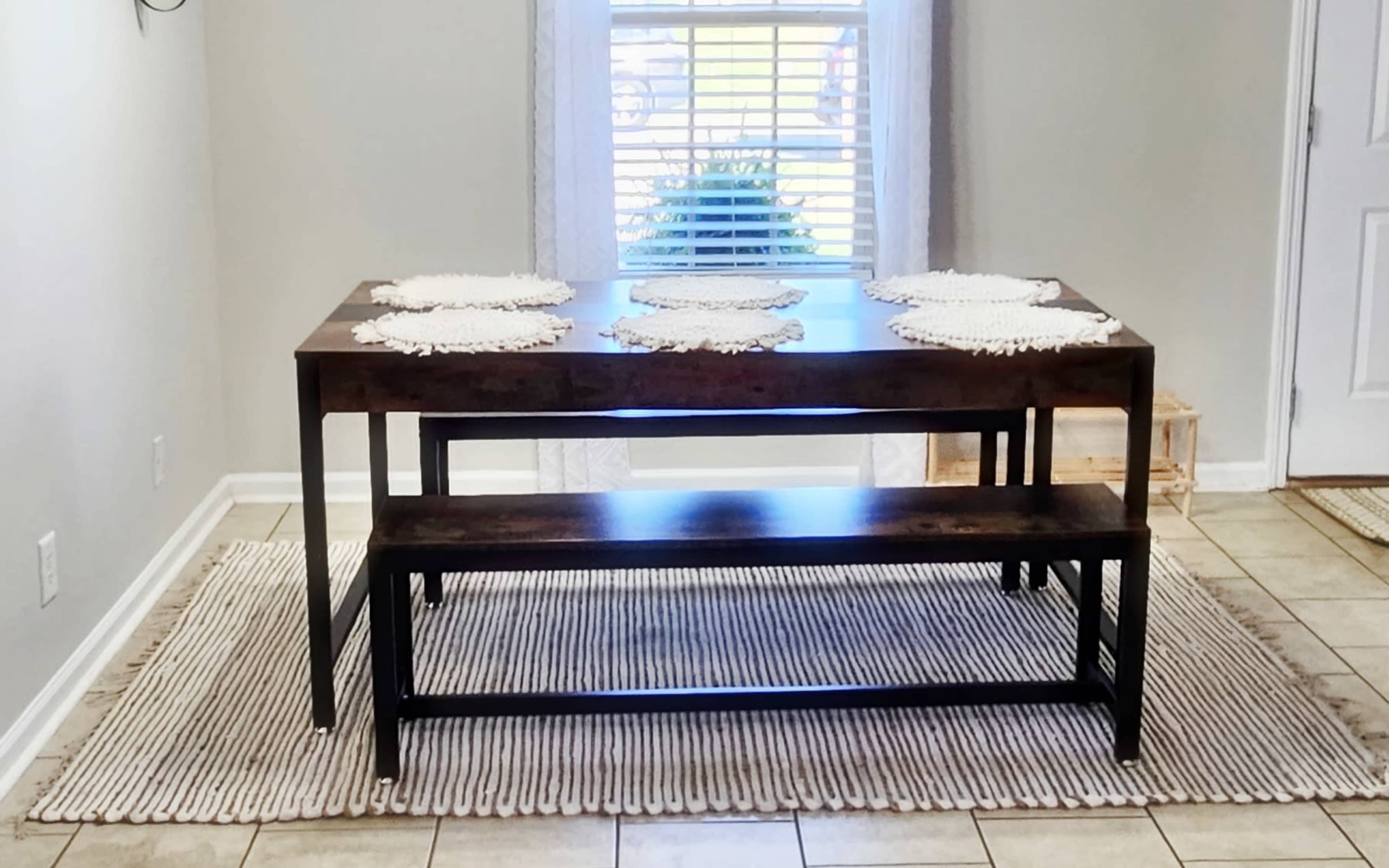 A rectangular wooden dining table with a bench sits on a striped rug in a well-lit room, with a window covered by a sheer curtain in the background.