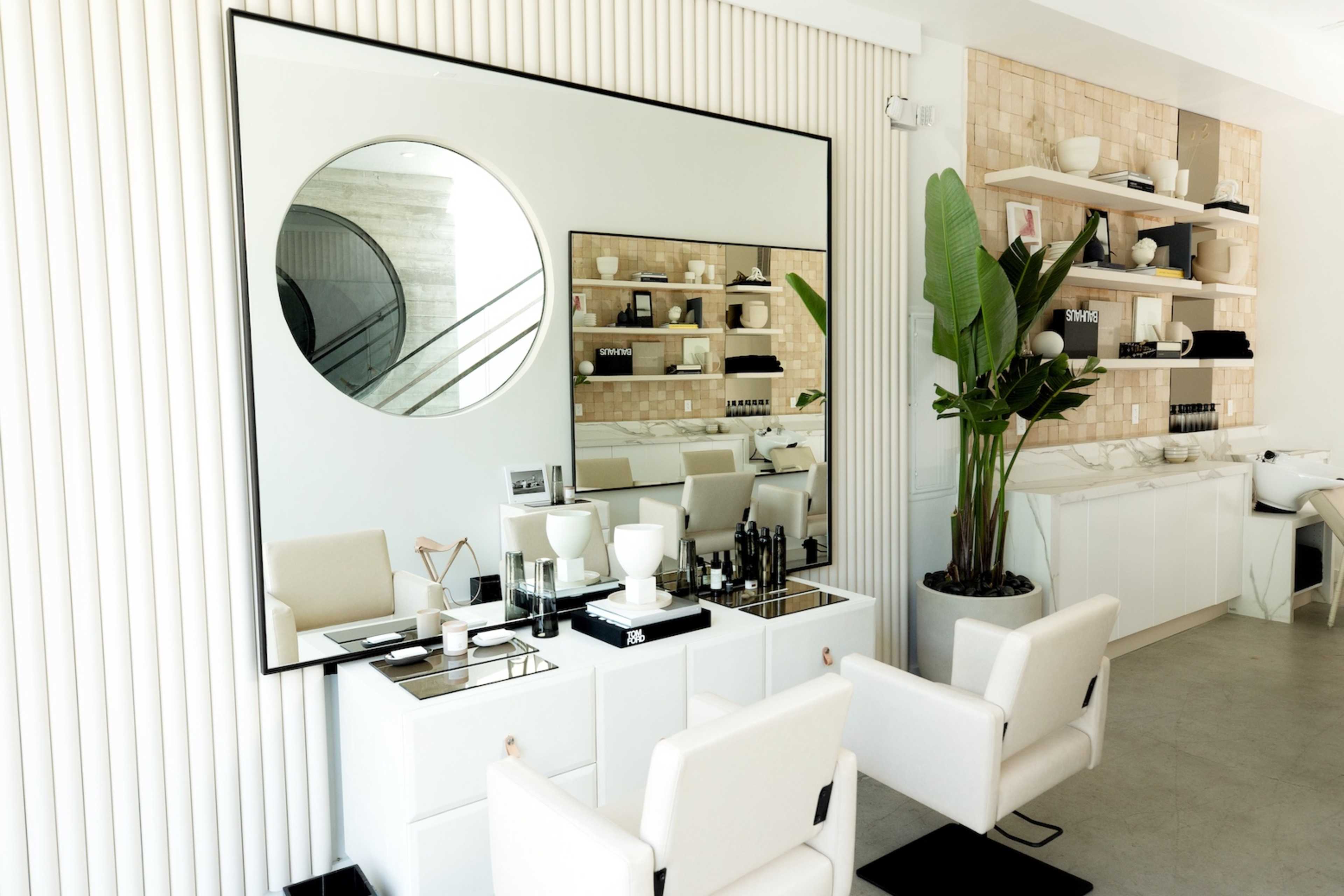 A modern hair salon with a large circular mirror, minimalist furniture, and neatly arranged products on a shelf in a bright, open space.