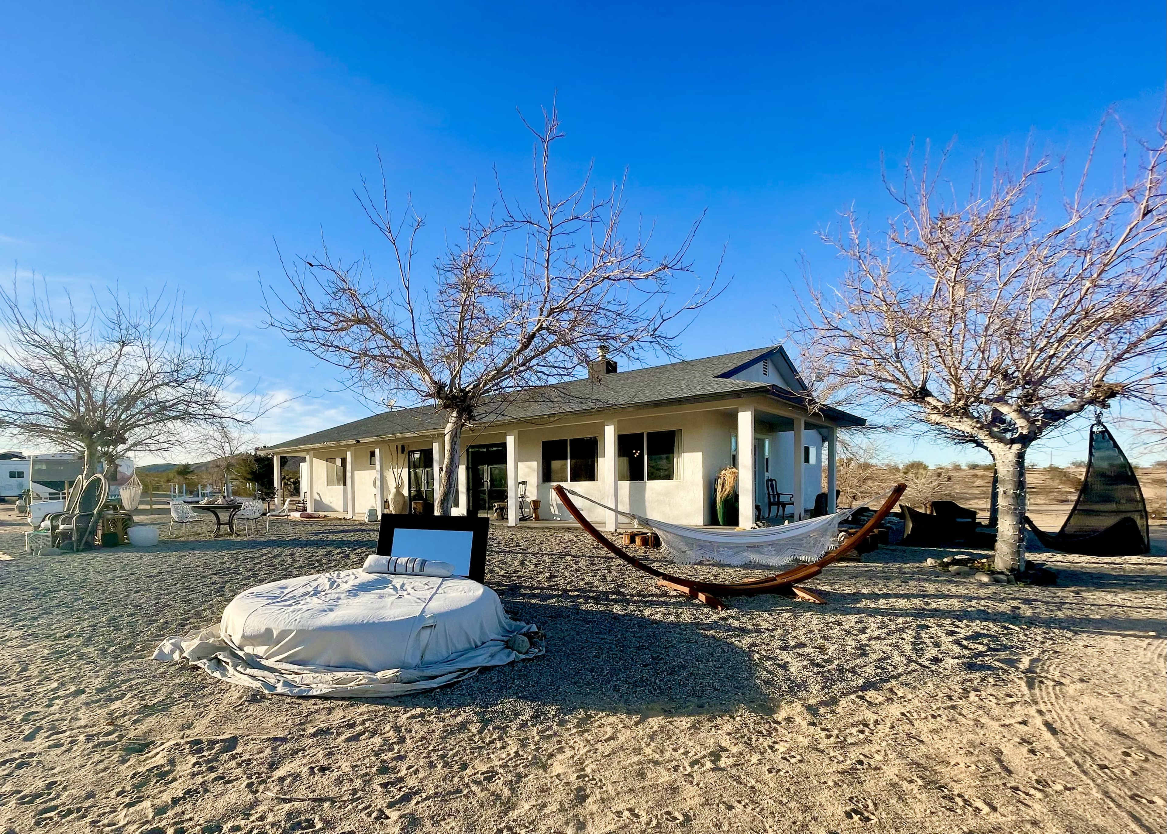 A desert property features a house surrounded by bare trees, outdoor seating, and hammocks on a sandy area.