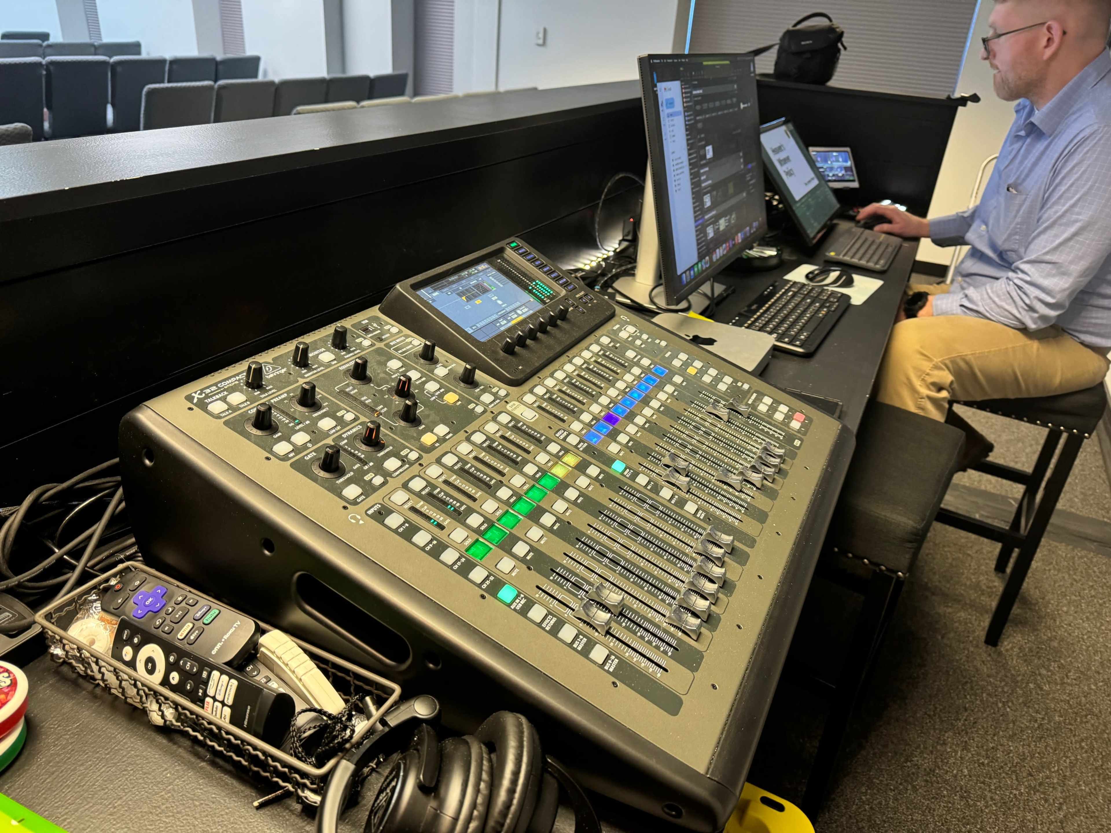 A sound mixing console is positioned on a table next to a laptop and desktop computer in a spacious room with chairs in the background.