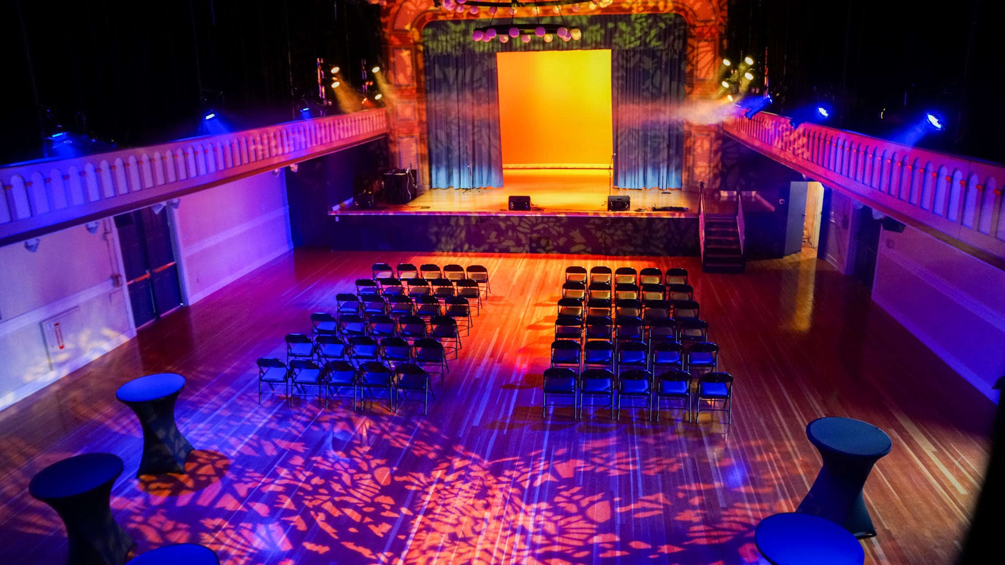 The image shows a large performance space with rows of black chairs arranged in front of a stage, illuminated by colorful lights.