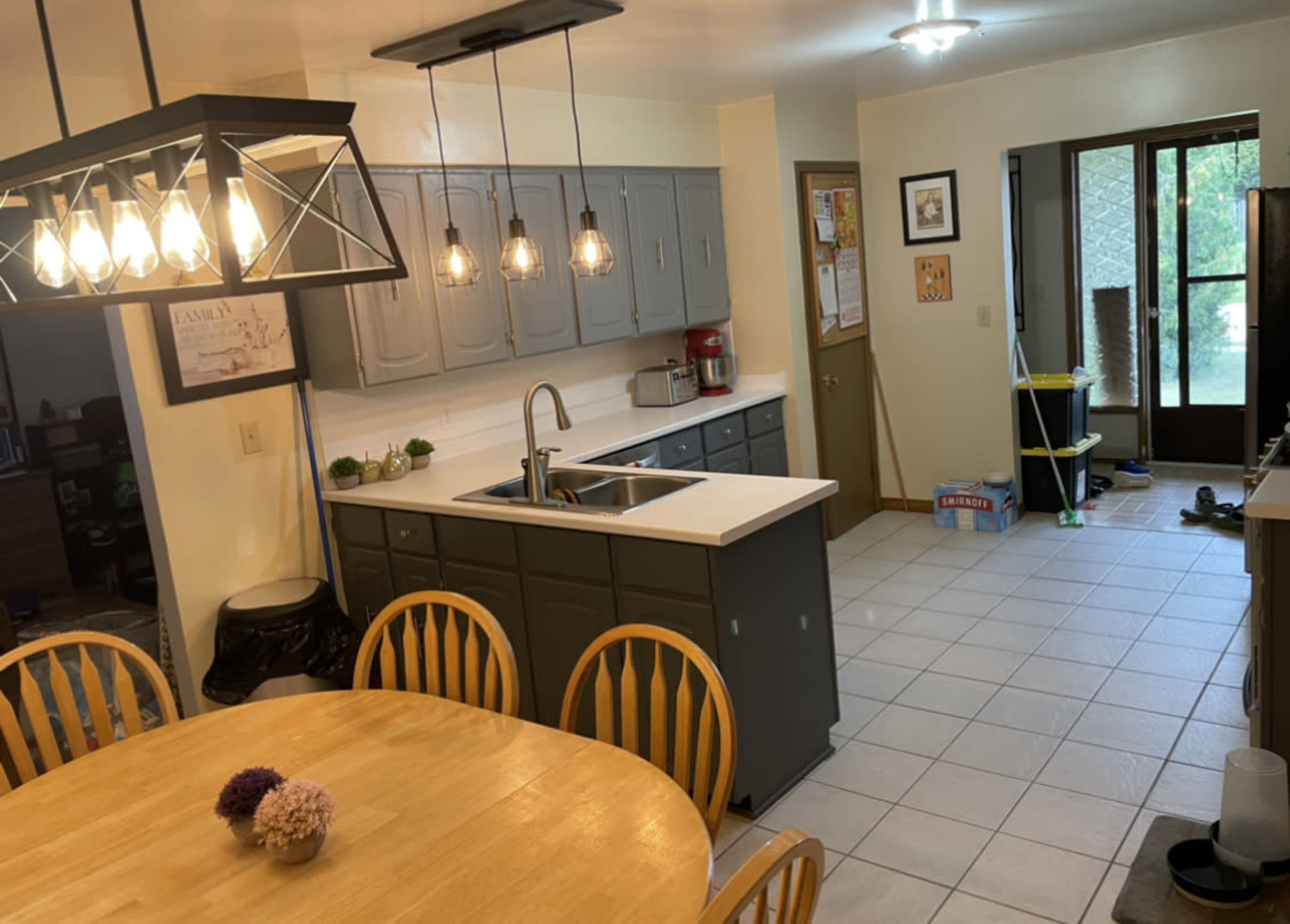 A bright kitchen with gray cabinets, a central island with a sink, a wooden dining table, and a doorway leading to another room.