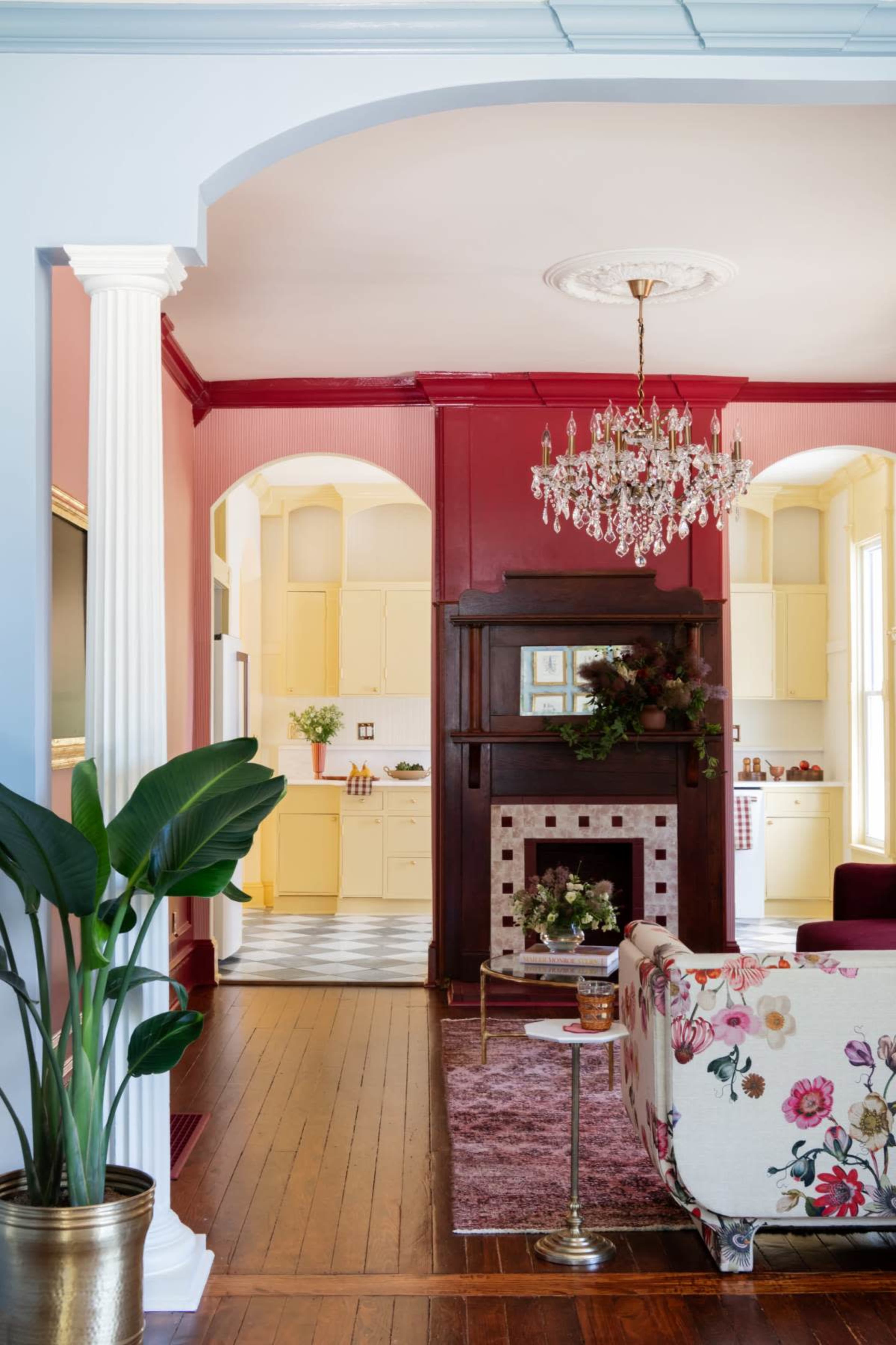 The image shows a bright living room that opens into a kitchen, featuring a floral-patterned sofa, a chandelier, and a fireplace with decorative elements.