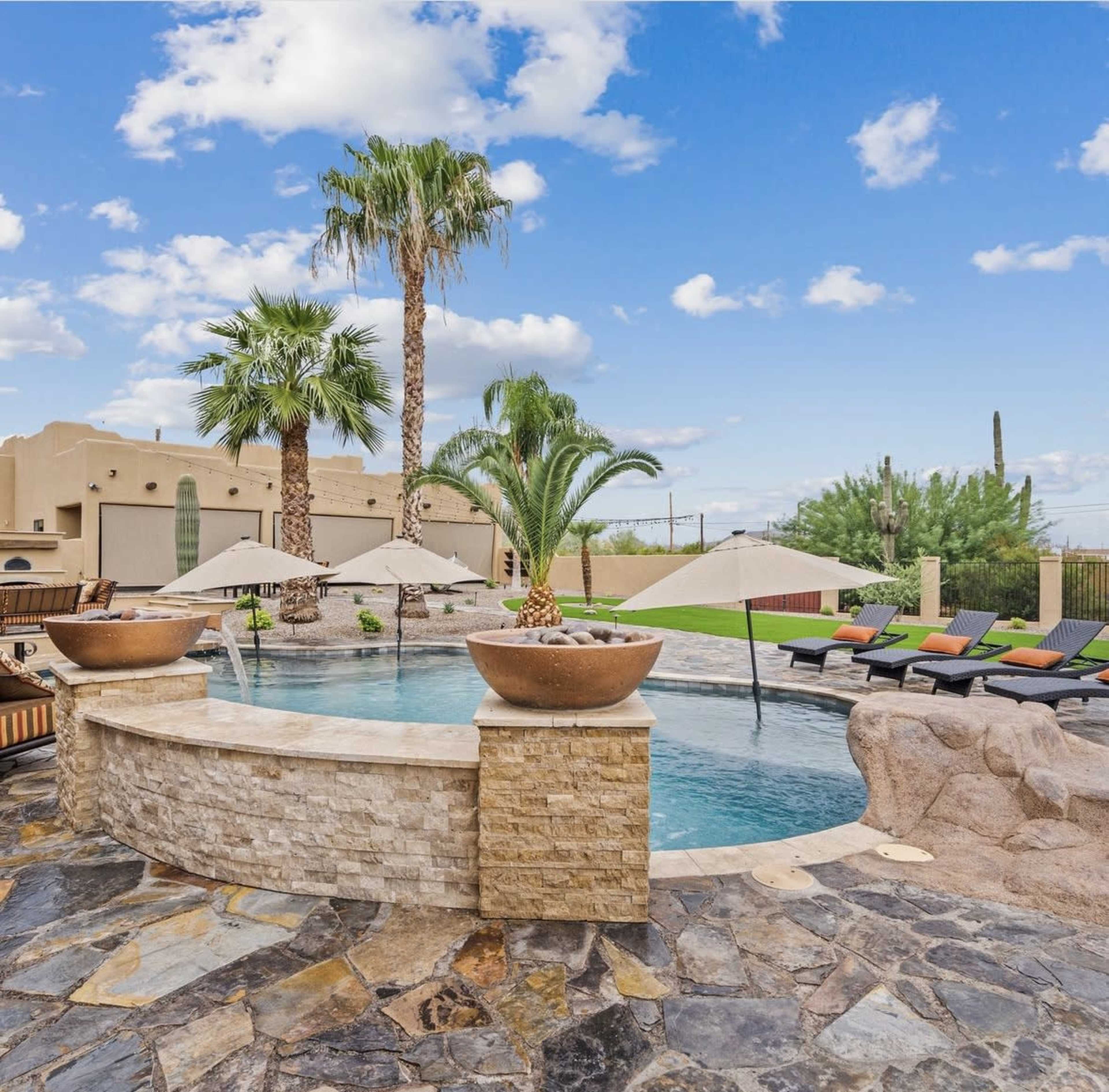 The image shows a backyard pool surrounded by palm trees and lounge chairs, with water features cascading into the pool.