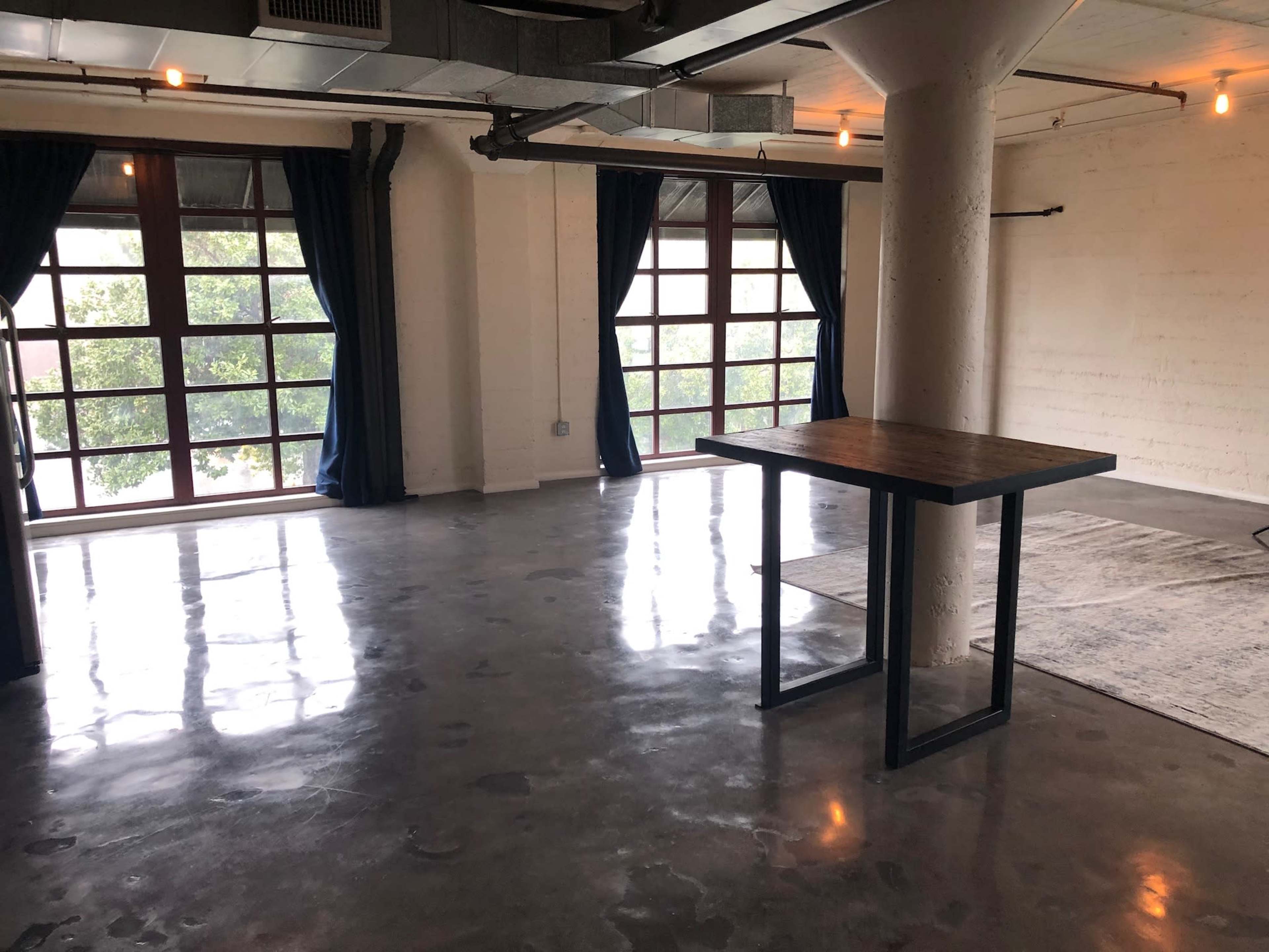 A spacious, empty room with polished concrete floors, large windows, and a wooden table in the center.
