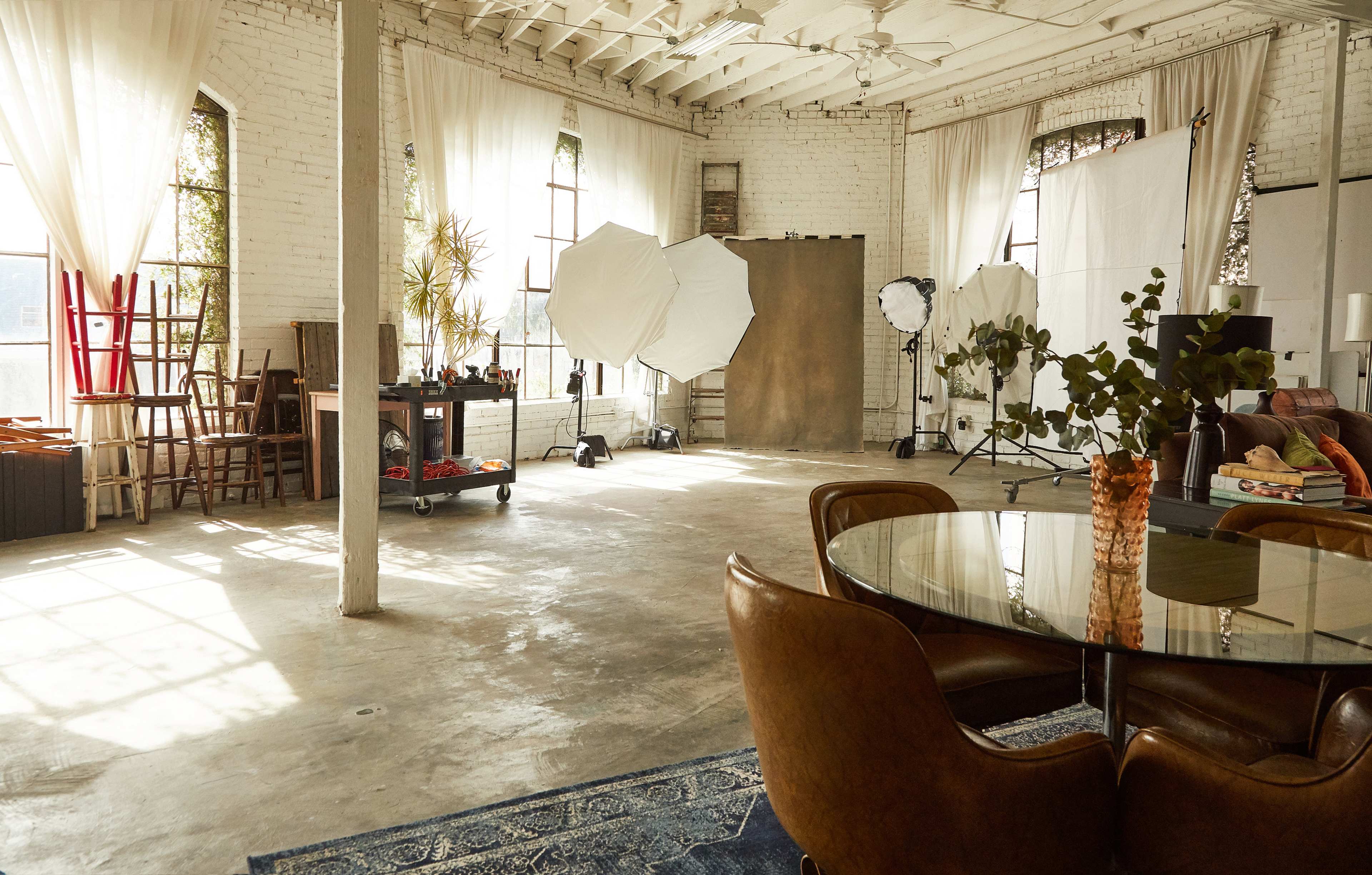 The image shows a spacious, well-lit studio with large windows, a dining table, and photography equipment set up in the background.