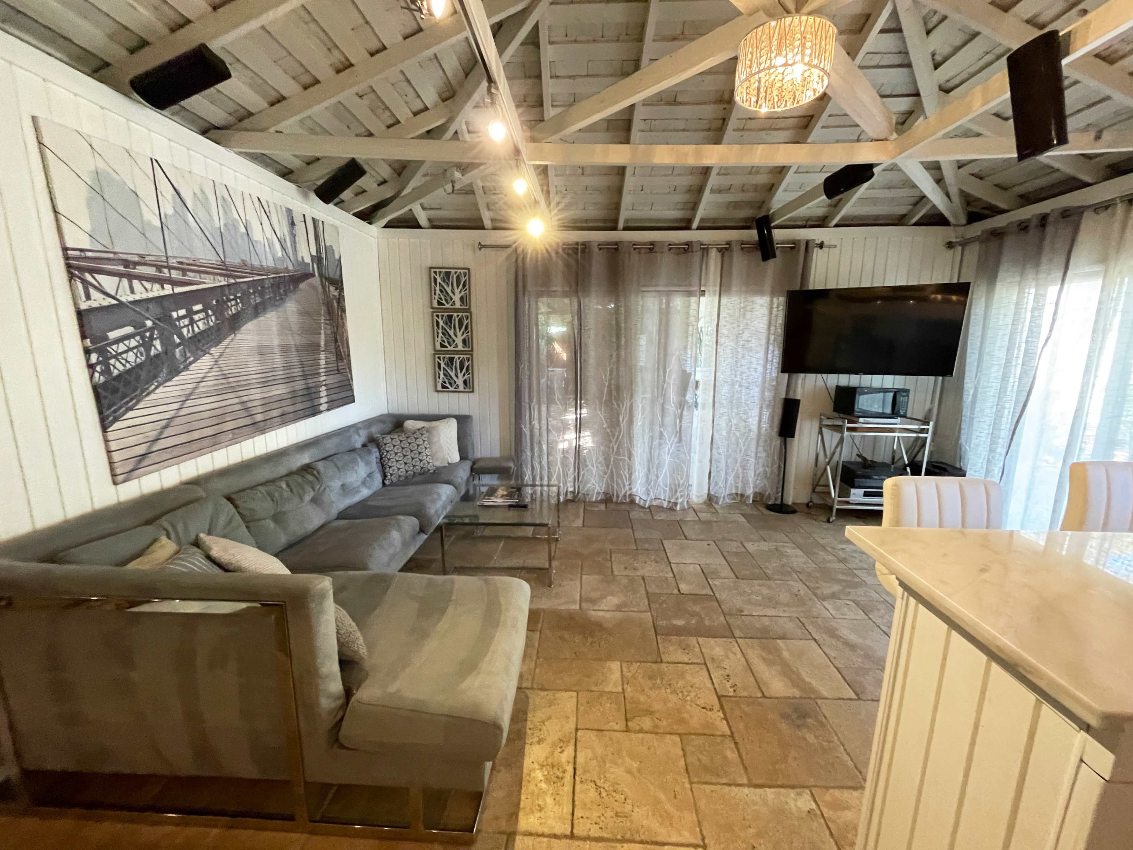 The image shows a cozy interior room with a sectional sofa, a mounted television, and large windows covered by sheer curtains.