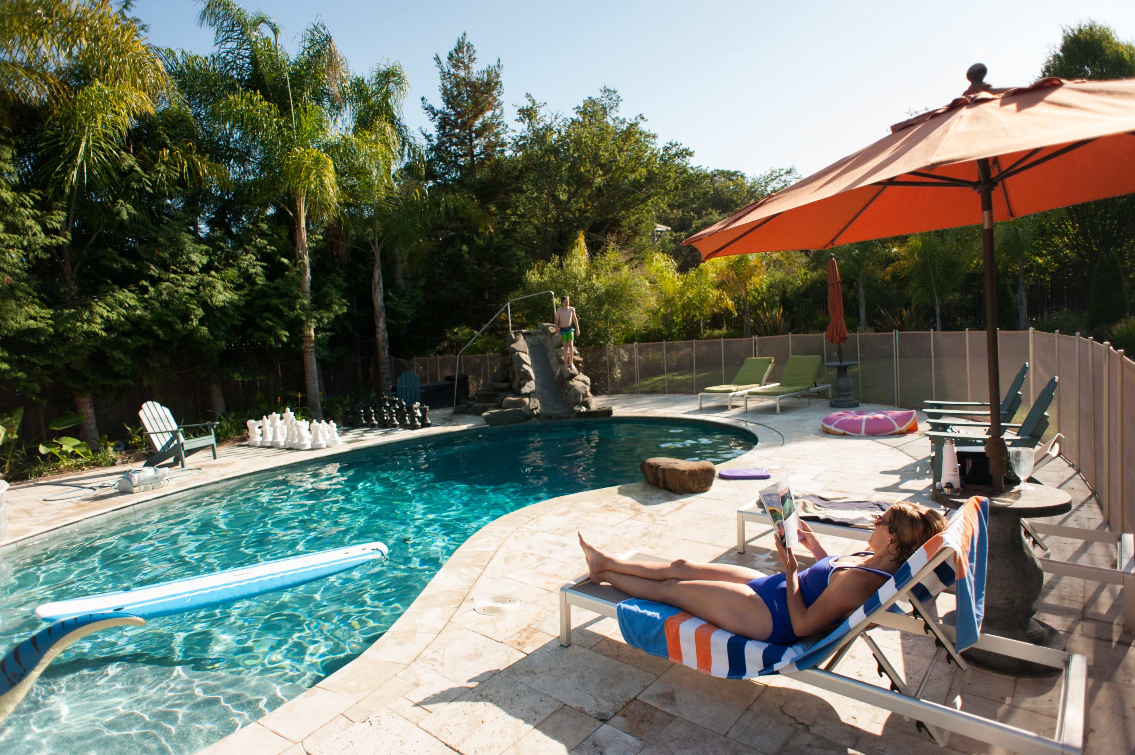 A woman relaxes on a lounge chair beside a swimming pool with palm trees and a rock formation in the background.