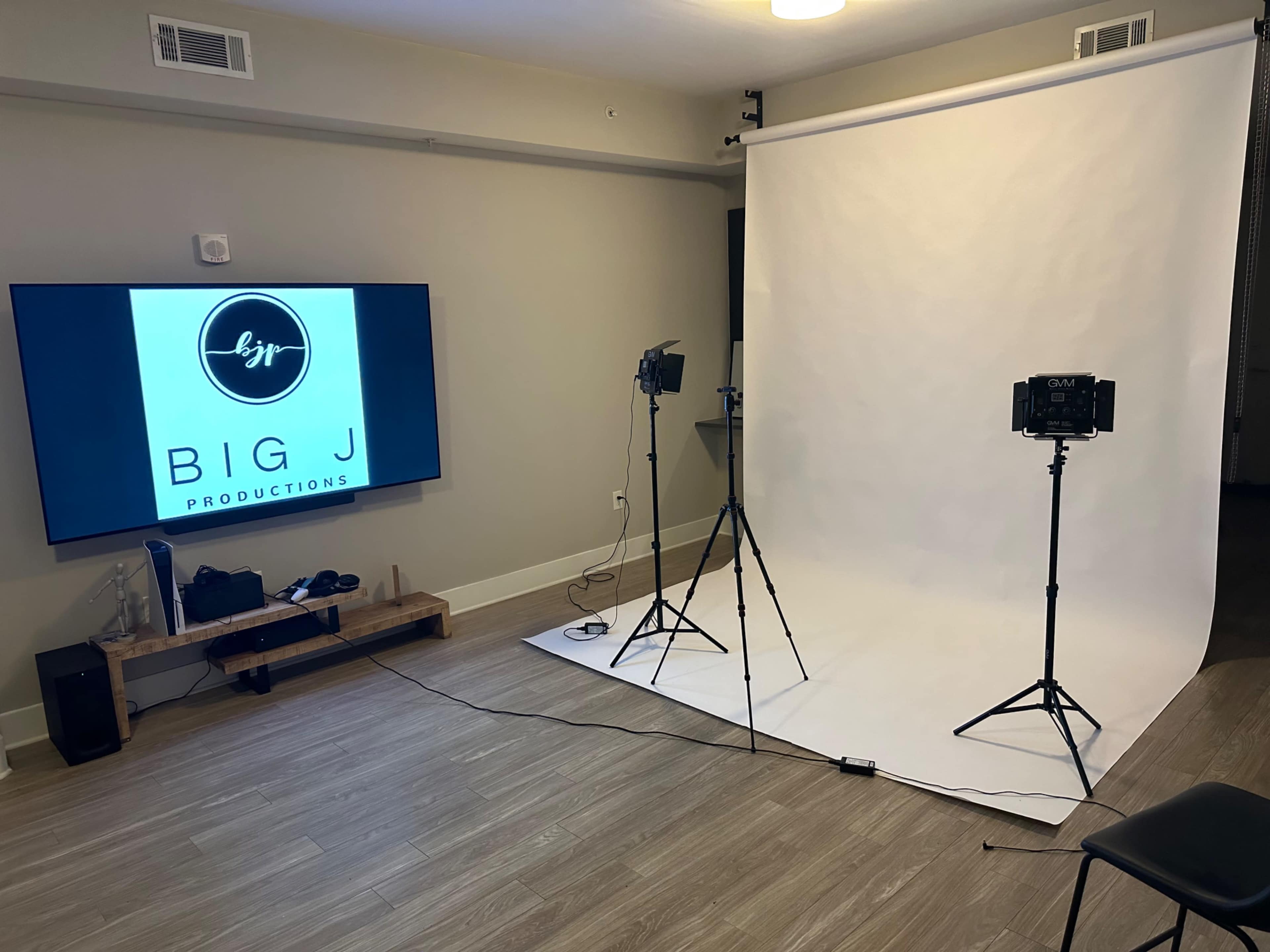 A photography setup with a white backdrop, two tripods holding lights, and a large screen displaying "BIG J Productions" on the wall.