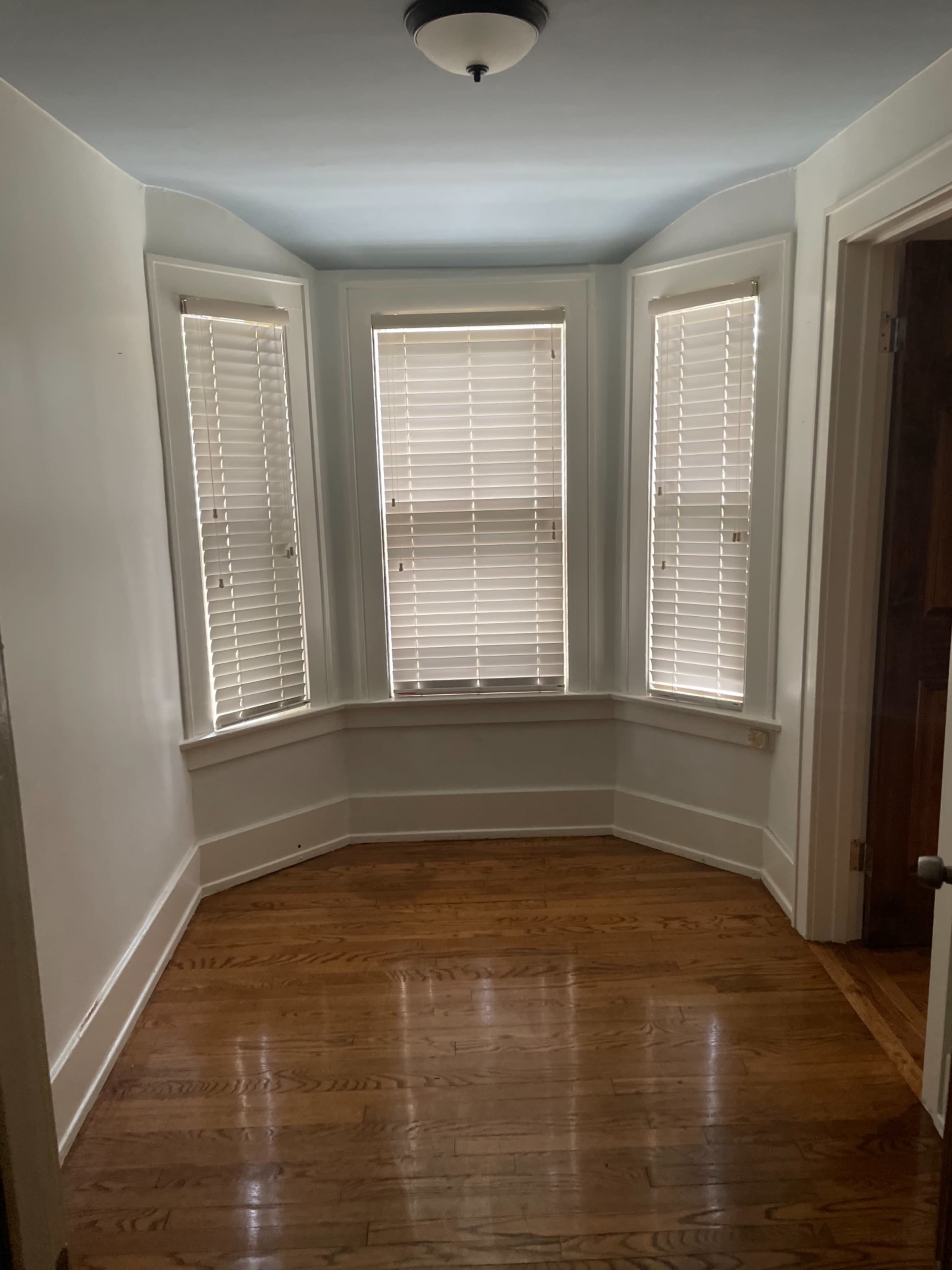 A small room with three windows featuring blinds, hardwood flooring, and a light fixture on the ceiling.
