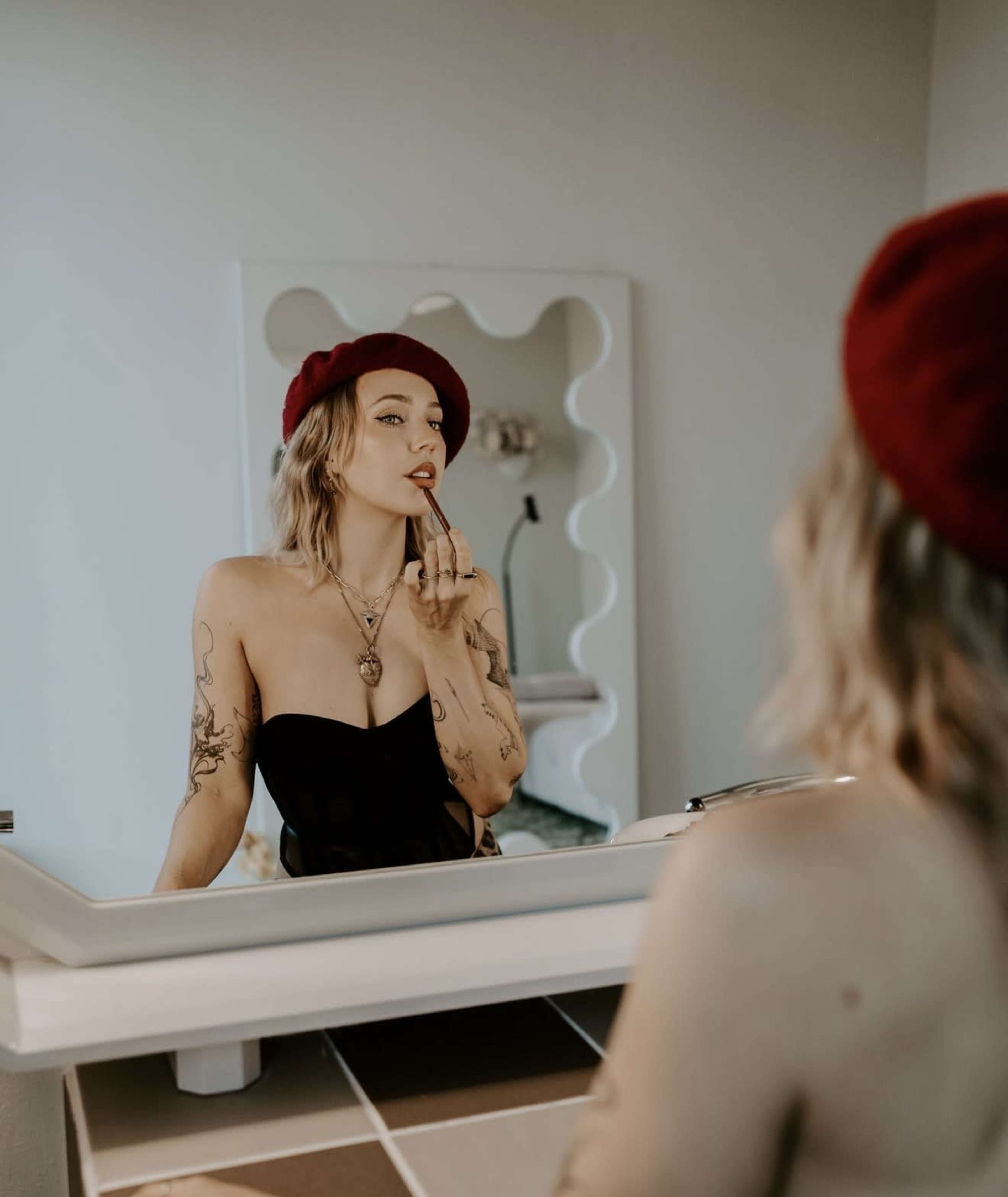A woman with tattoos is applying lipstick while looking into a mirror, wearing a black top and a red beret.