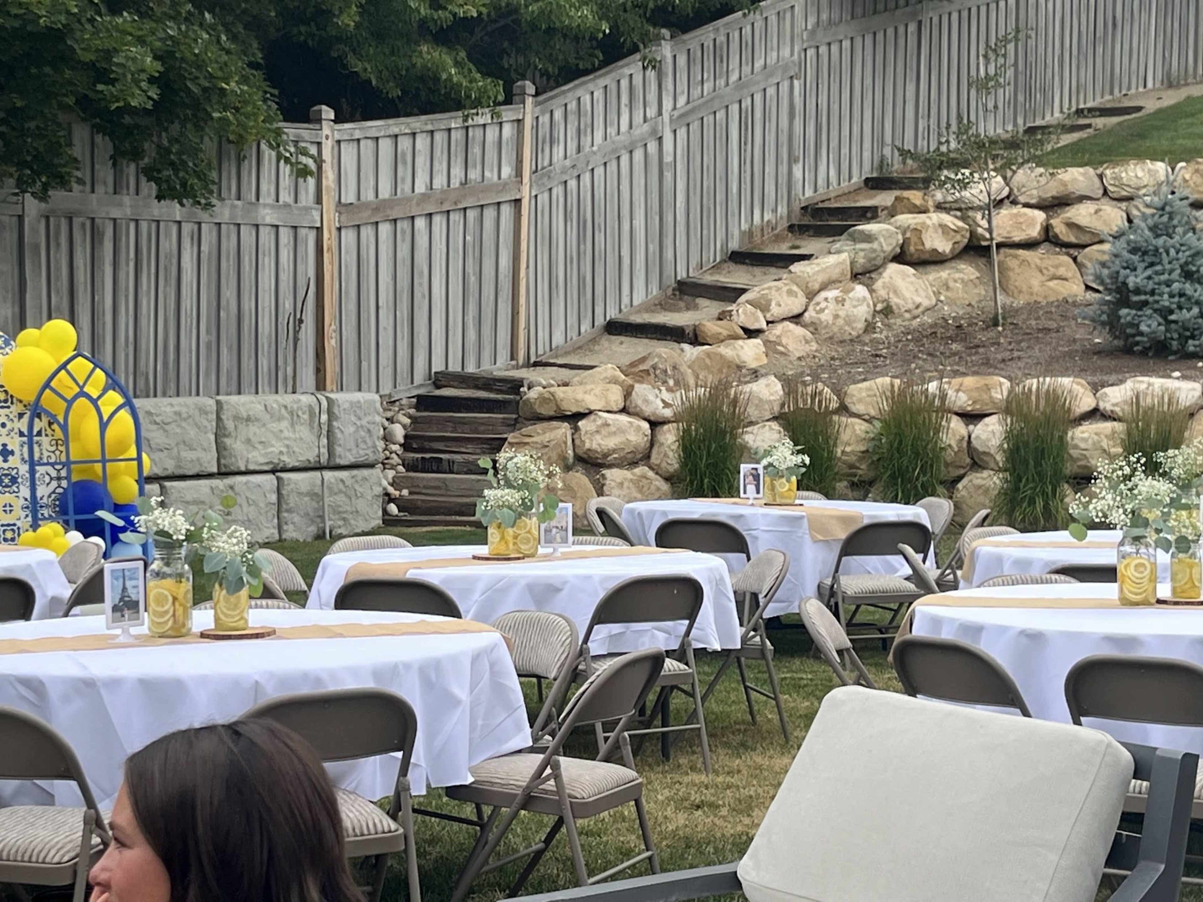 The image shows a backyard setting for an event, featuring several round tables covered with white tablecloths, arranged on a lawn with a wooden fence and stone wall in the background.