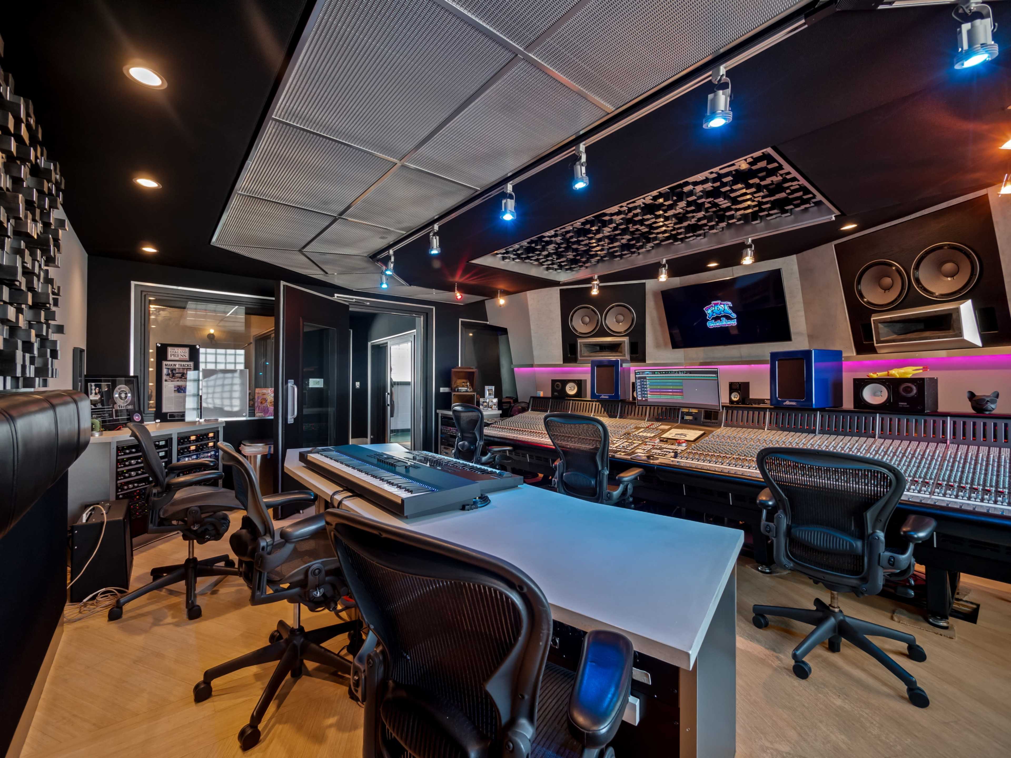 The image shows a modern music studio equipped with a large mixing console, multiple speakers, and soundproofing panels.