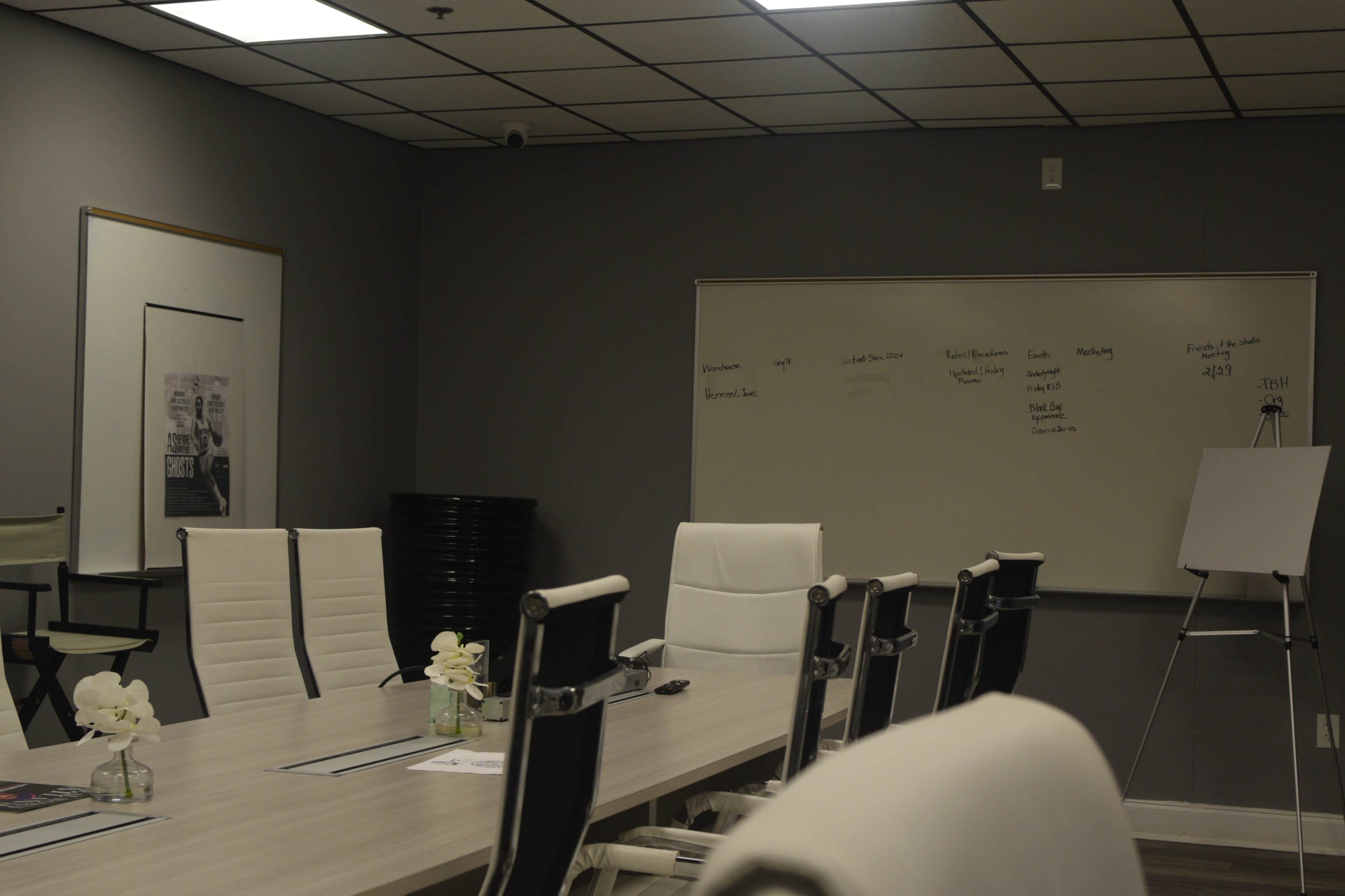 A modern conference room features a long table surrounded by white chairs, a whiteboard with handwritten notes, and a large poster on the wall.