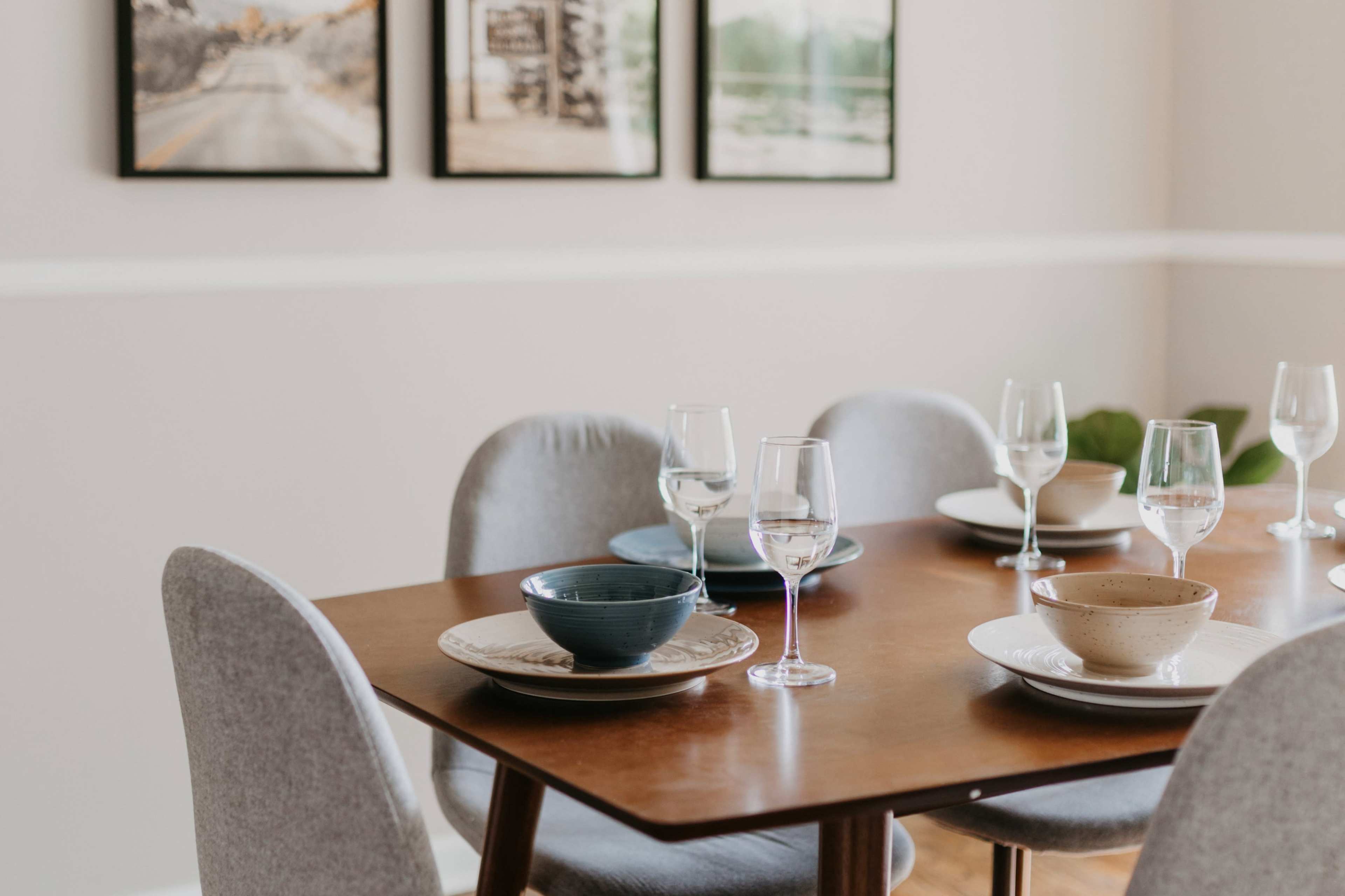 A dining table is set with plates and glassware, surrounded by chairs in a light-colored room with framed photographs on the wall.