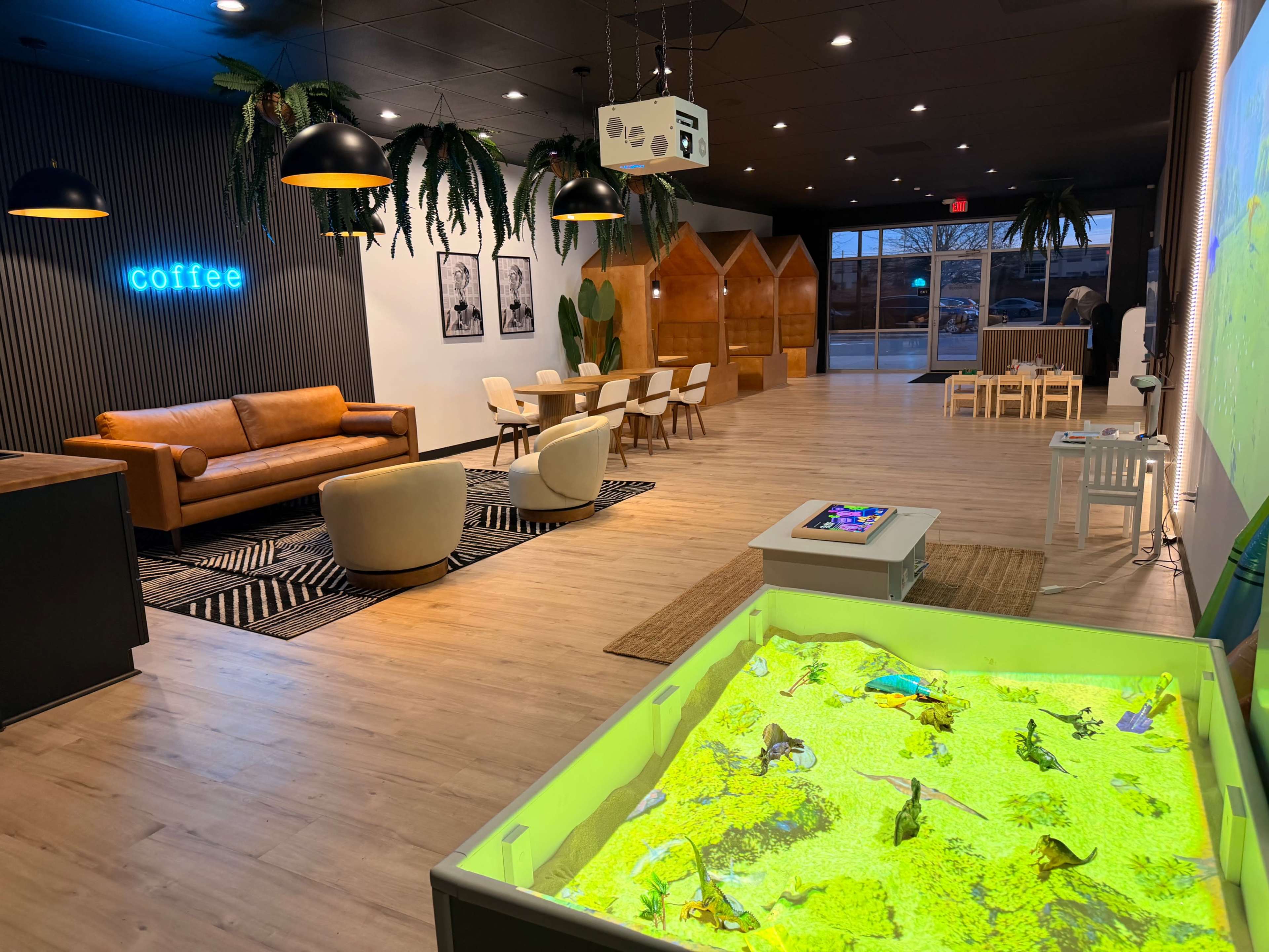The image depicts a modern indoor space featuring a coffee area with a couch, seating arrangements, and an interactive sandbox with bright green sand.