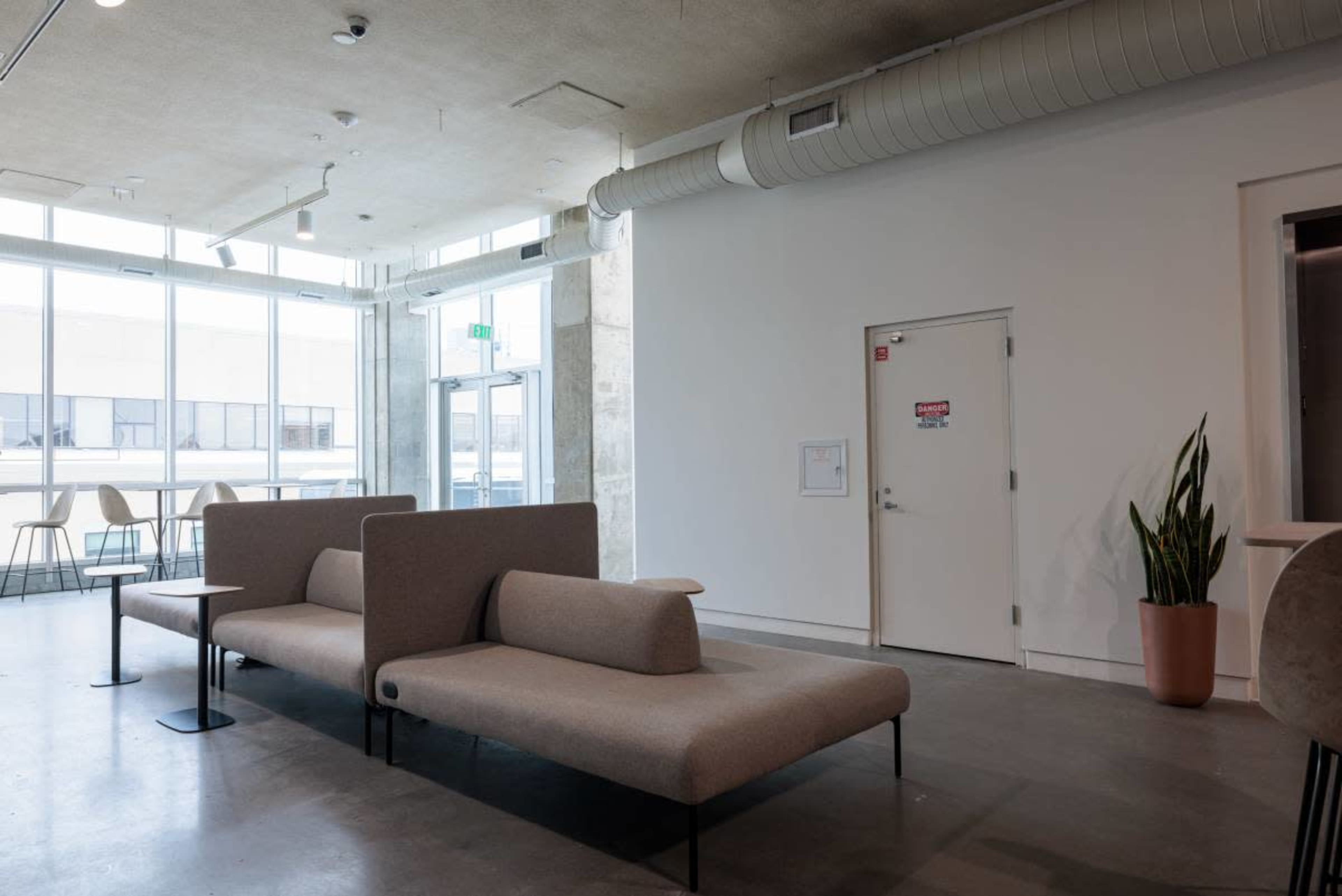 The image shows a modern lounge area with minimalist furniture, including sofas and a potted plant, adjacent to large windows and a door.