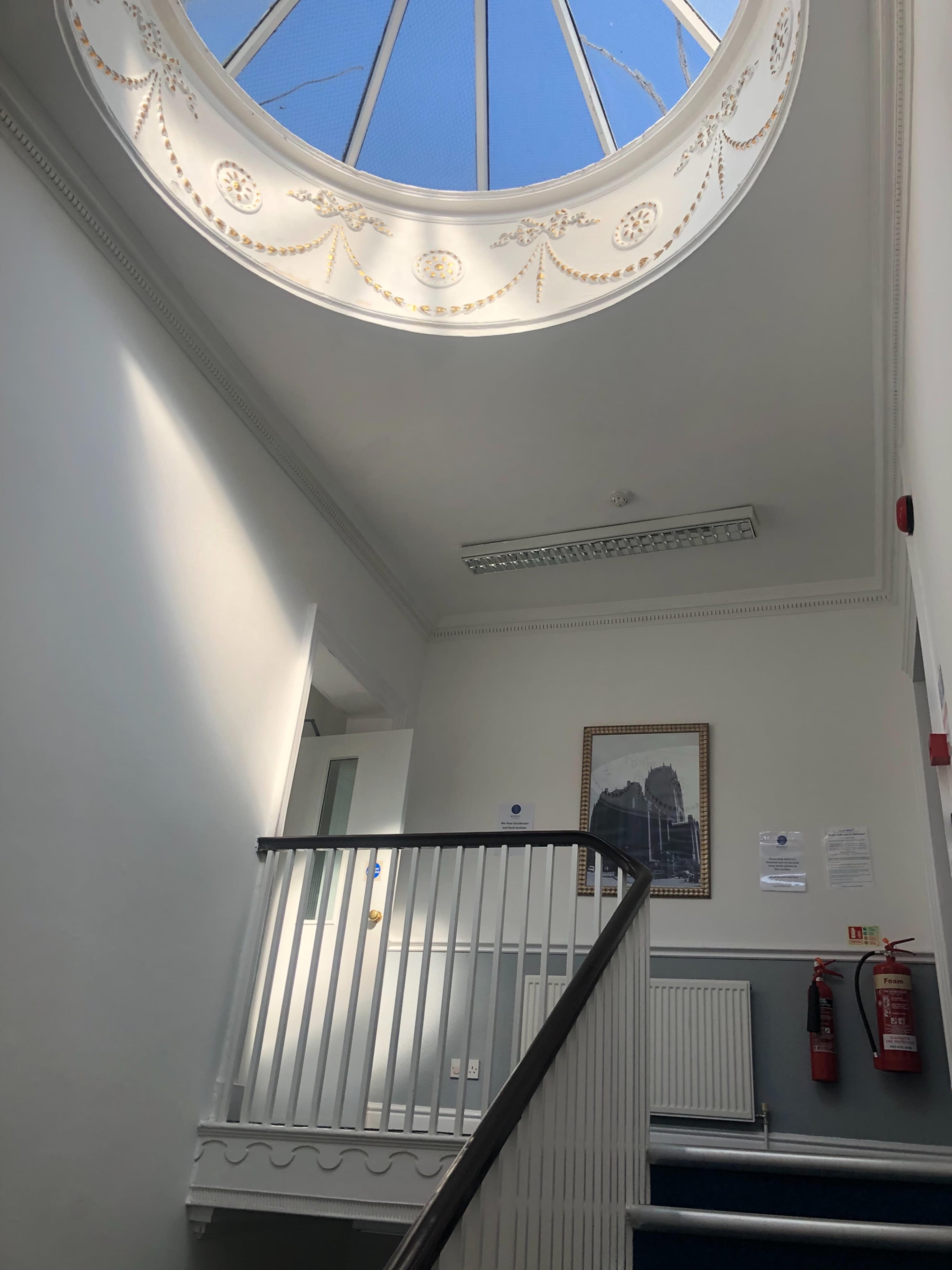 The image shows a stairwell with a circular skylight above, featuring a white railing and framed artwork on the wall.