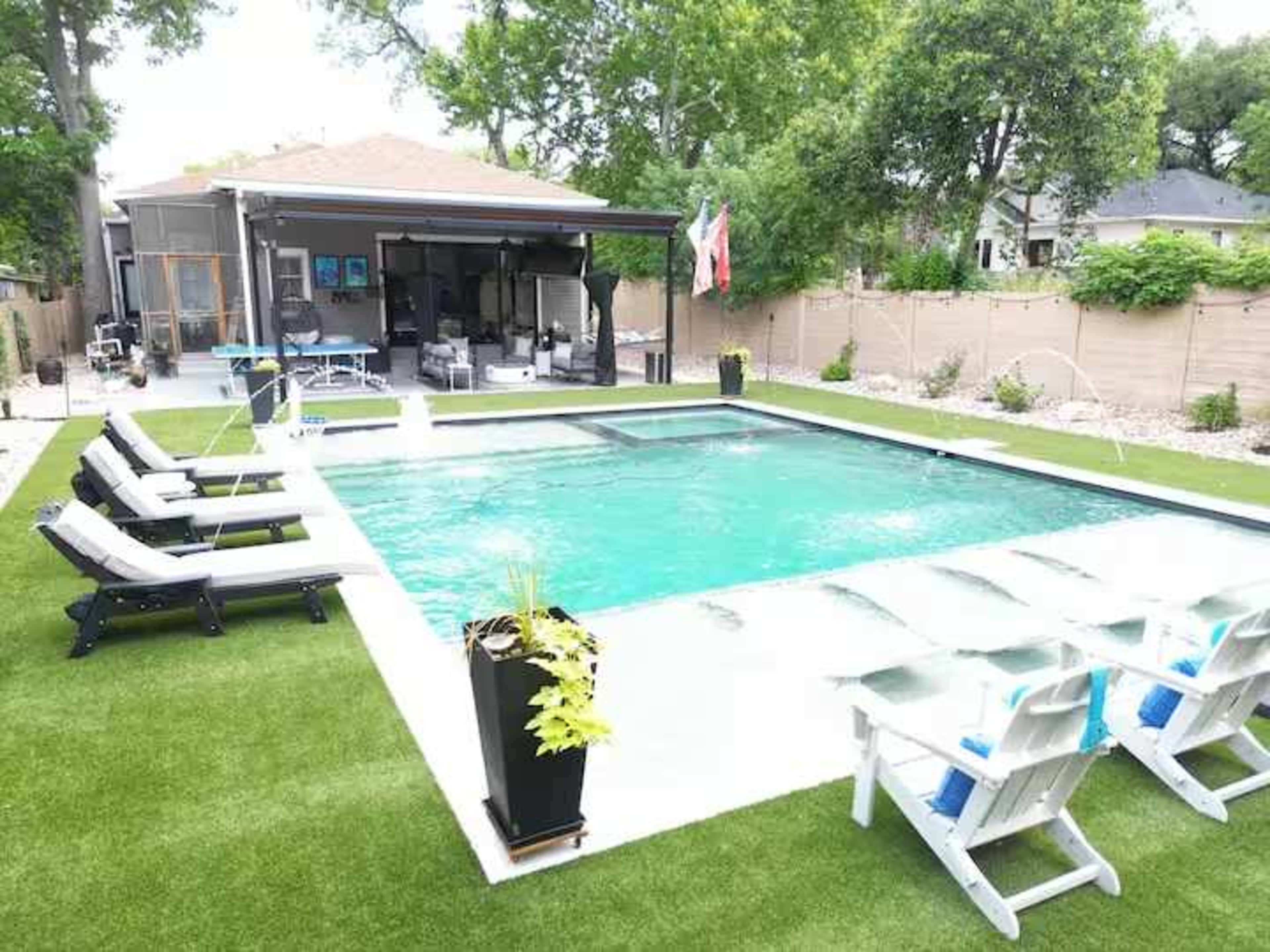 A rectangular swimming pool surrounded by lounge chairs and greenery, with a covered patio area in the background.