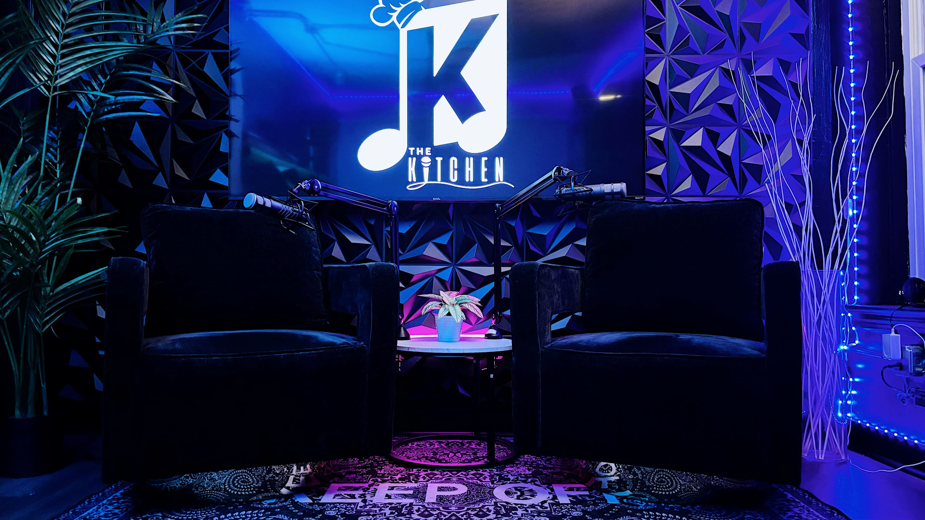 The image shows two black armchairs facing each other on a patterned rug in a studio environment, with a large screen displaying the text "The Kitchen" in the background.