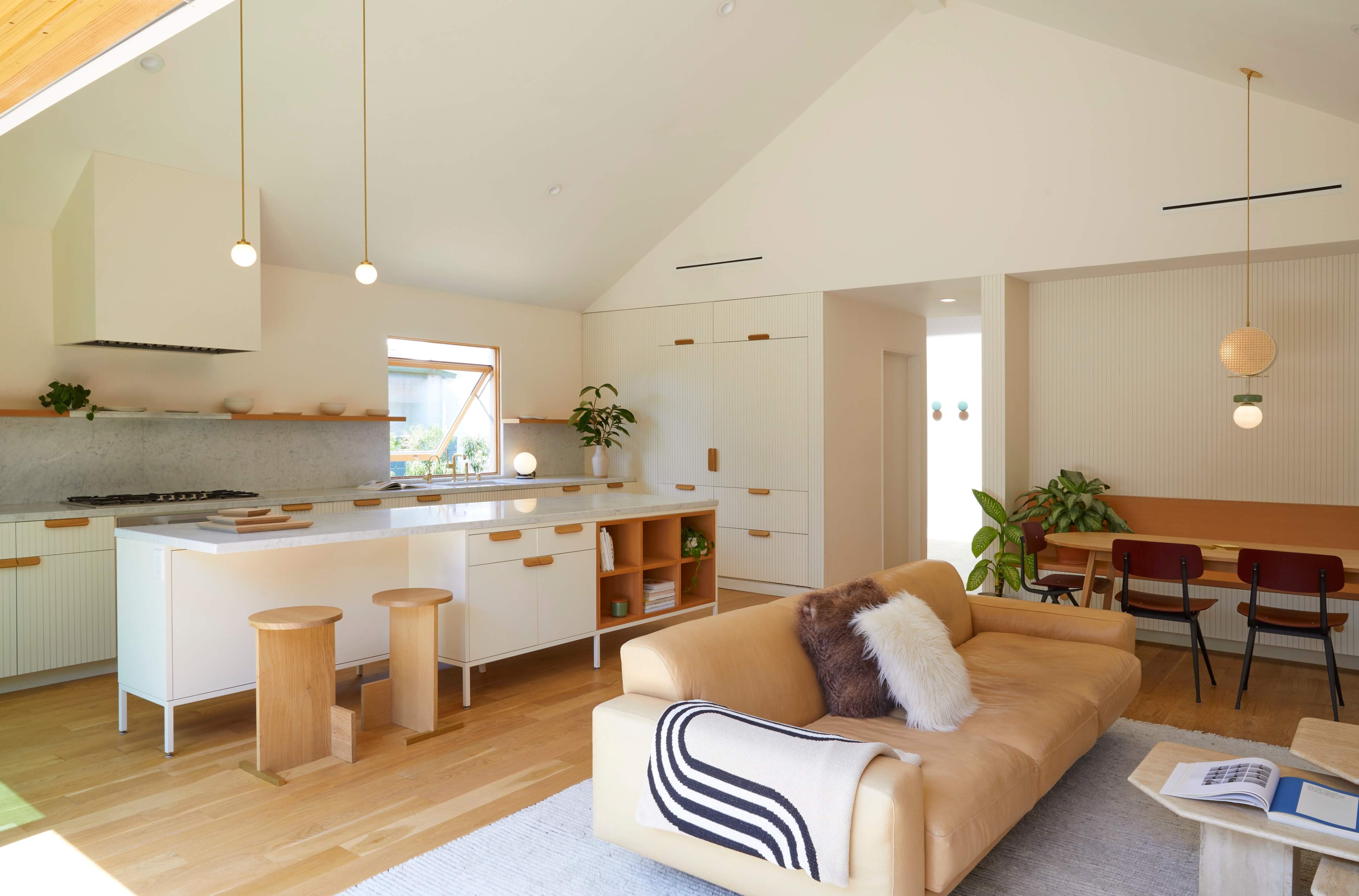 A modern kitchen and living area feature a minimalist design with a beige sofa, wooden stools, and large windows allowing natural light.