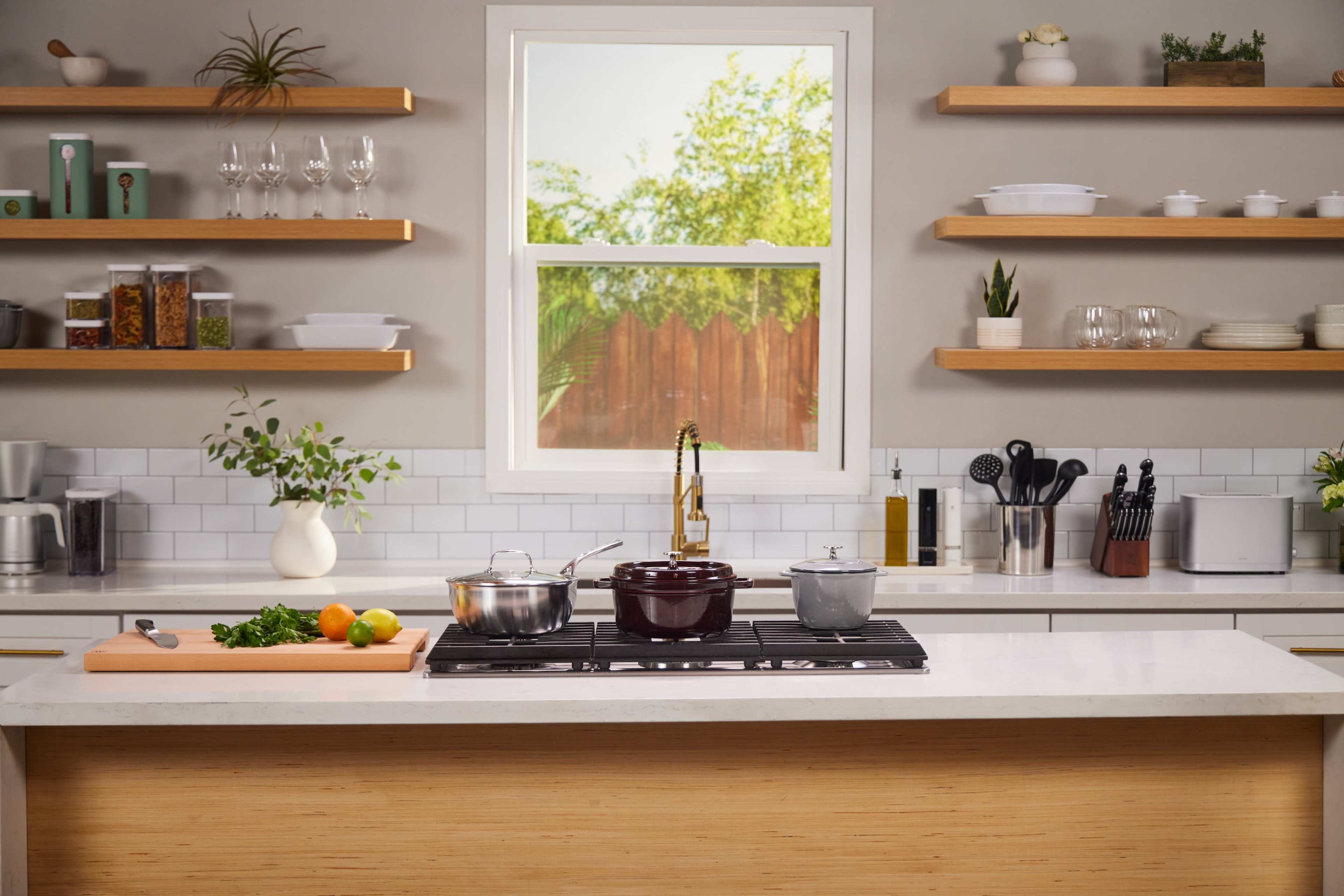A modern kitchen features a central island with cooking pots, a cutting board with vegetables, and open shelves displaying kitchenware and ingredients.