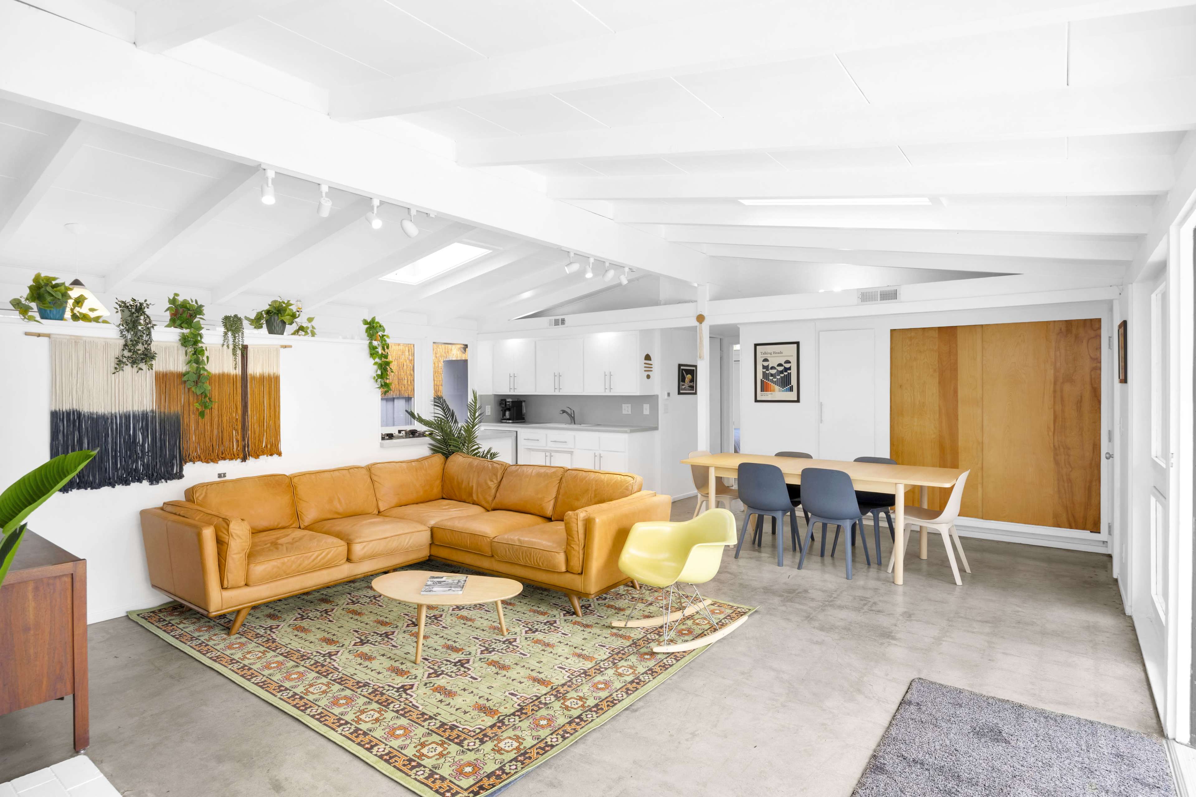 The image shows a bright, open living space featuring a tan sectional sofa, a wooden dining table, and a combination of plants and artwork on the walls.
