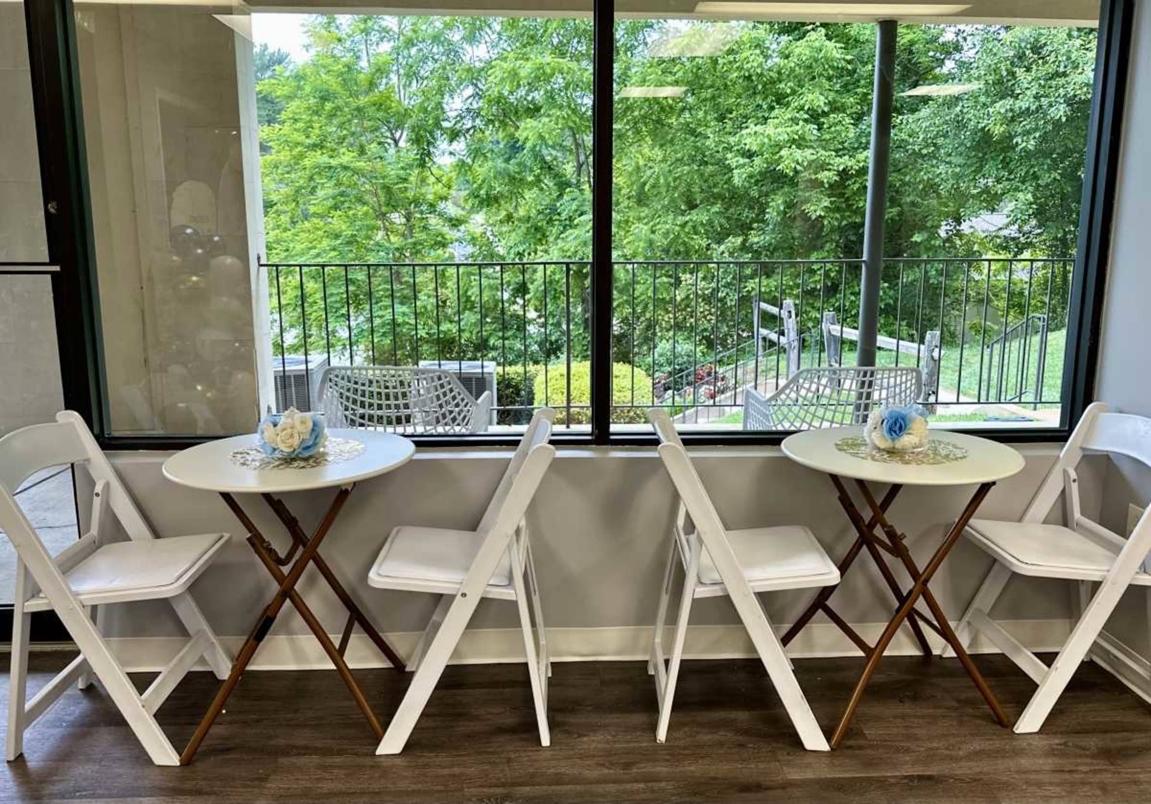 The image shows four white folding chairs arranged around two small round tables placed near a large window with greenery visible outside.