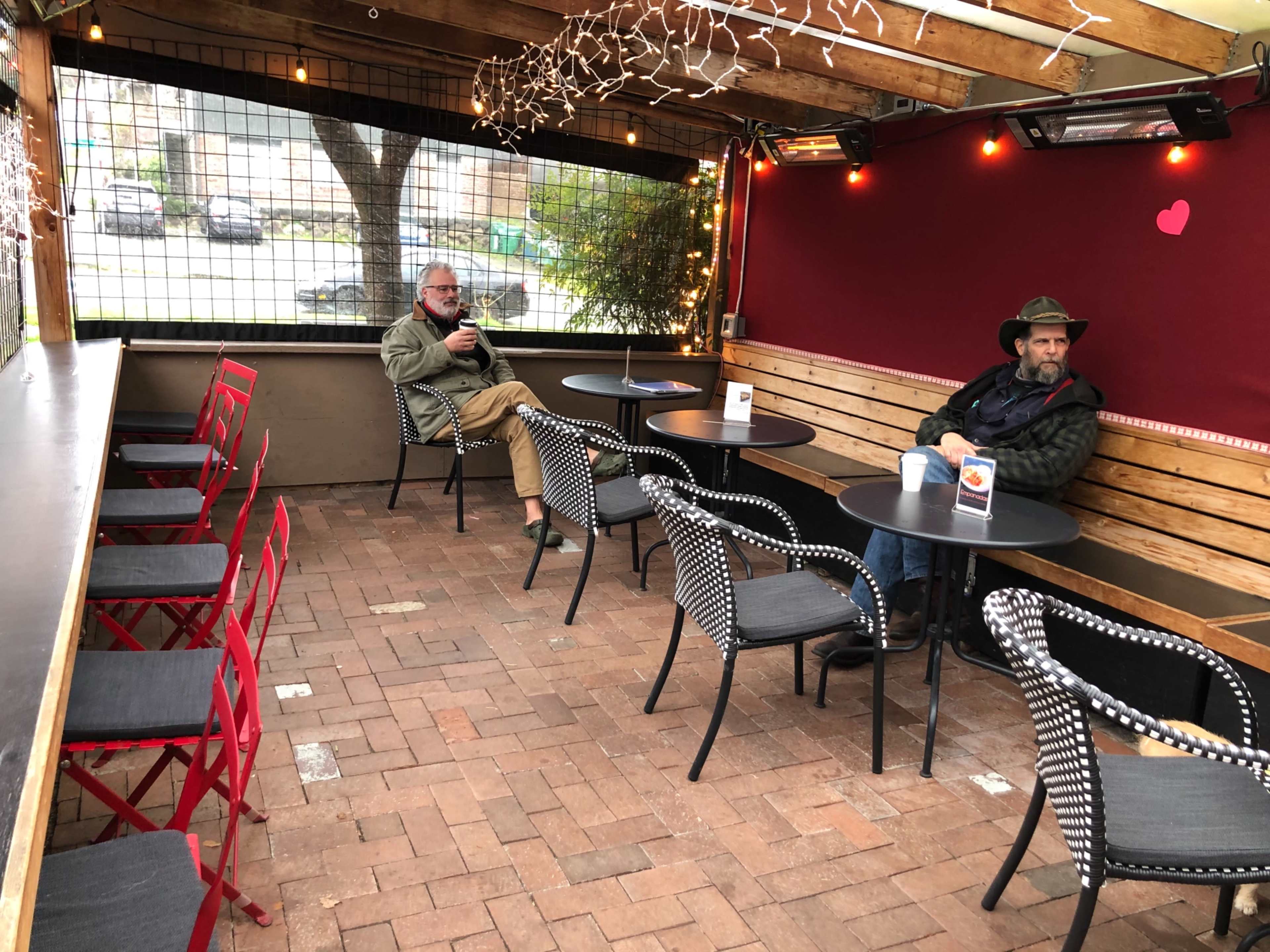 The image shows a partially enclosed café patio with several empty tables and two seated men, one in a hat and one in a jacket, against a backdrop of string lights.