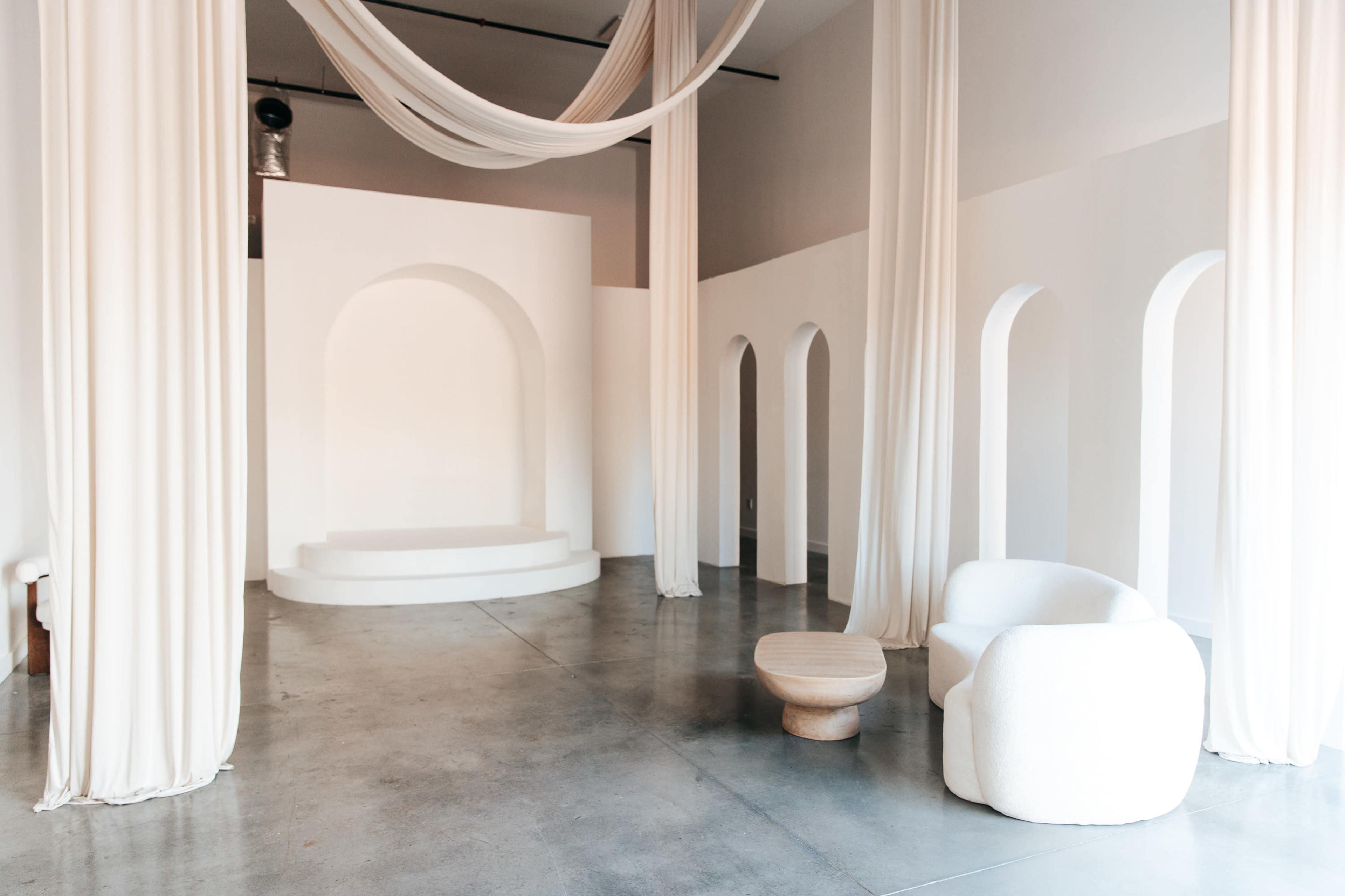 The image features a minimalist interior space with white walls, a raised platform, arched openings, and draped fabric.