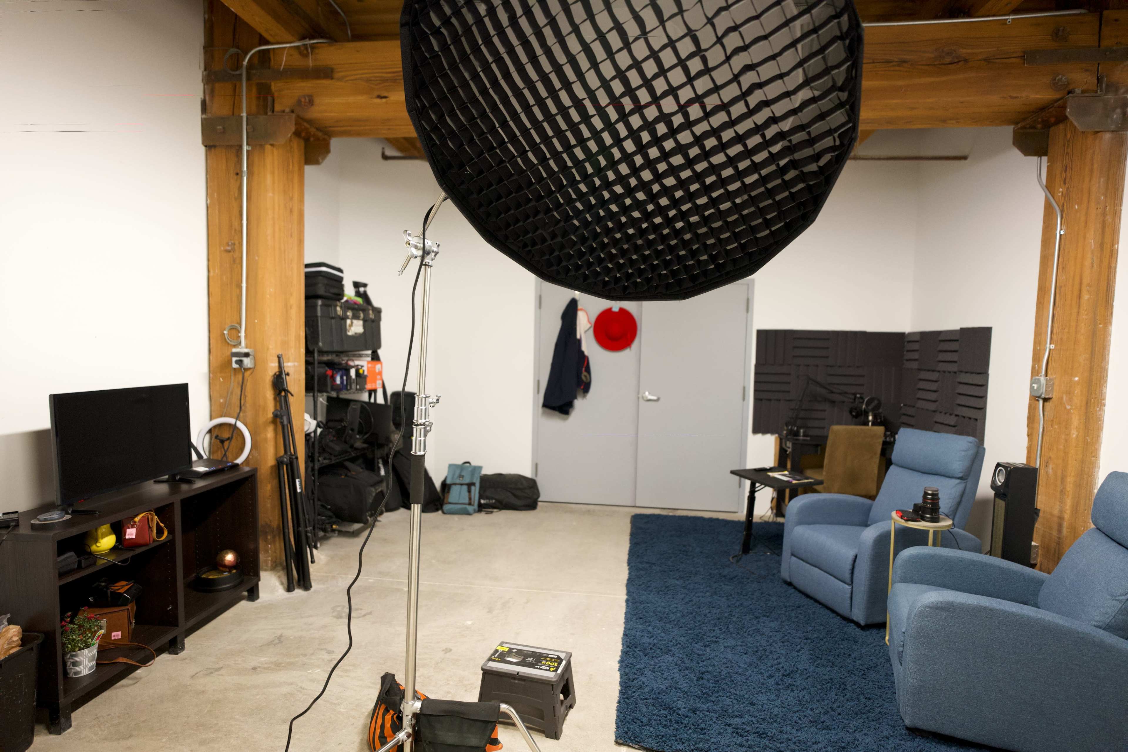 A spacious room with a blue rug, two blue chairs, a flat-screen TV on a stand, a lighting setup, and gray soundproofing panels on the wall.