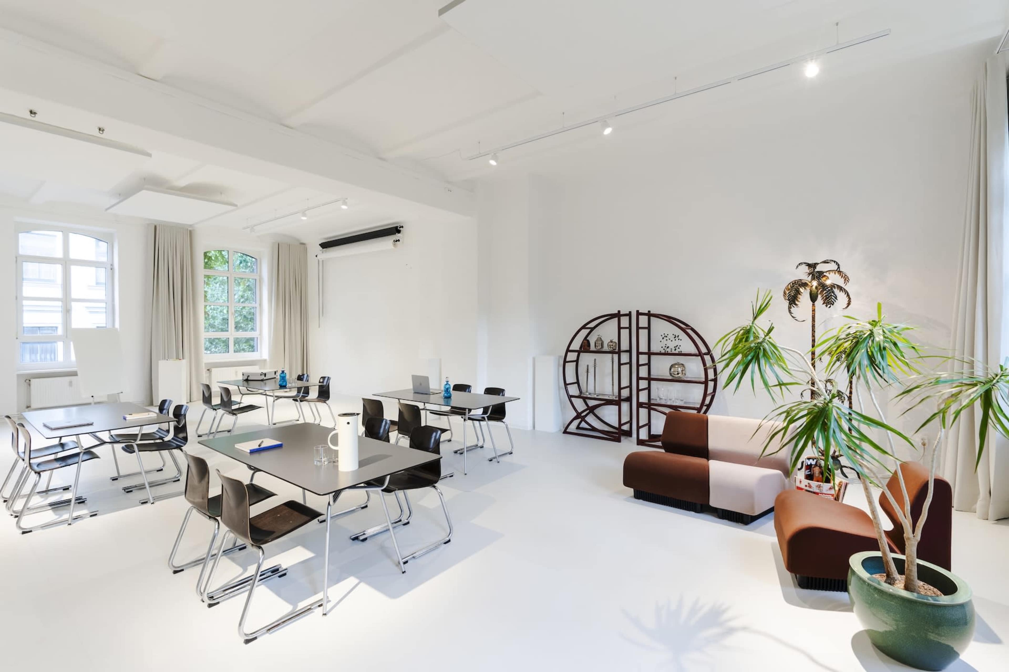 The image shows a bright, modern interior of a workspace with black tables, chairs, and a sofa, along with potted plants and large windows.
