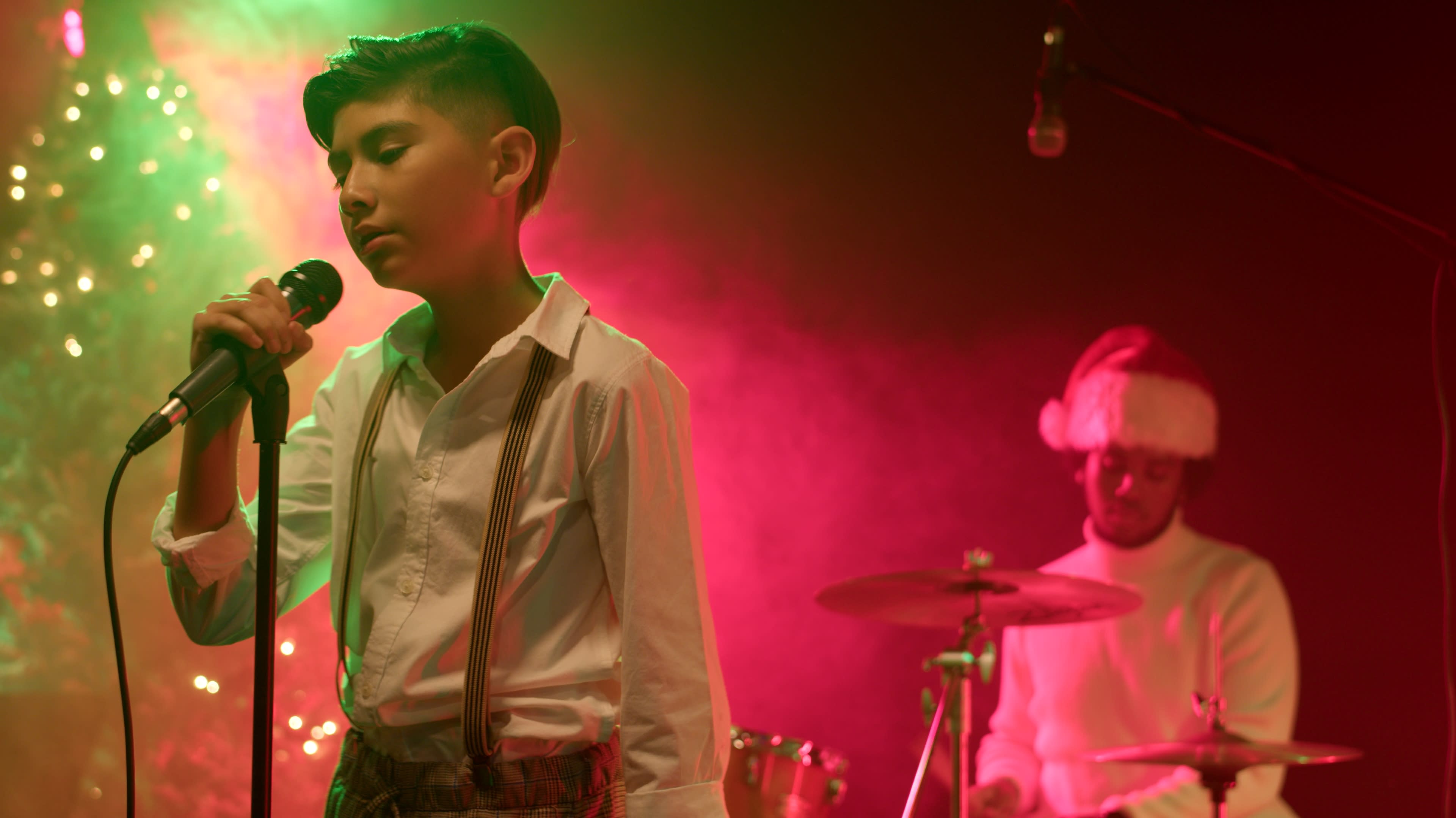 A young boy in a white shirt and suspenders sings into a microphone while a drummer in a Santa hat plays behind him in a colorful lighting setup.