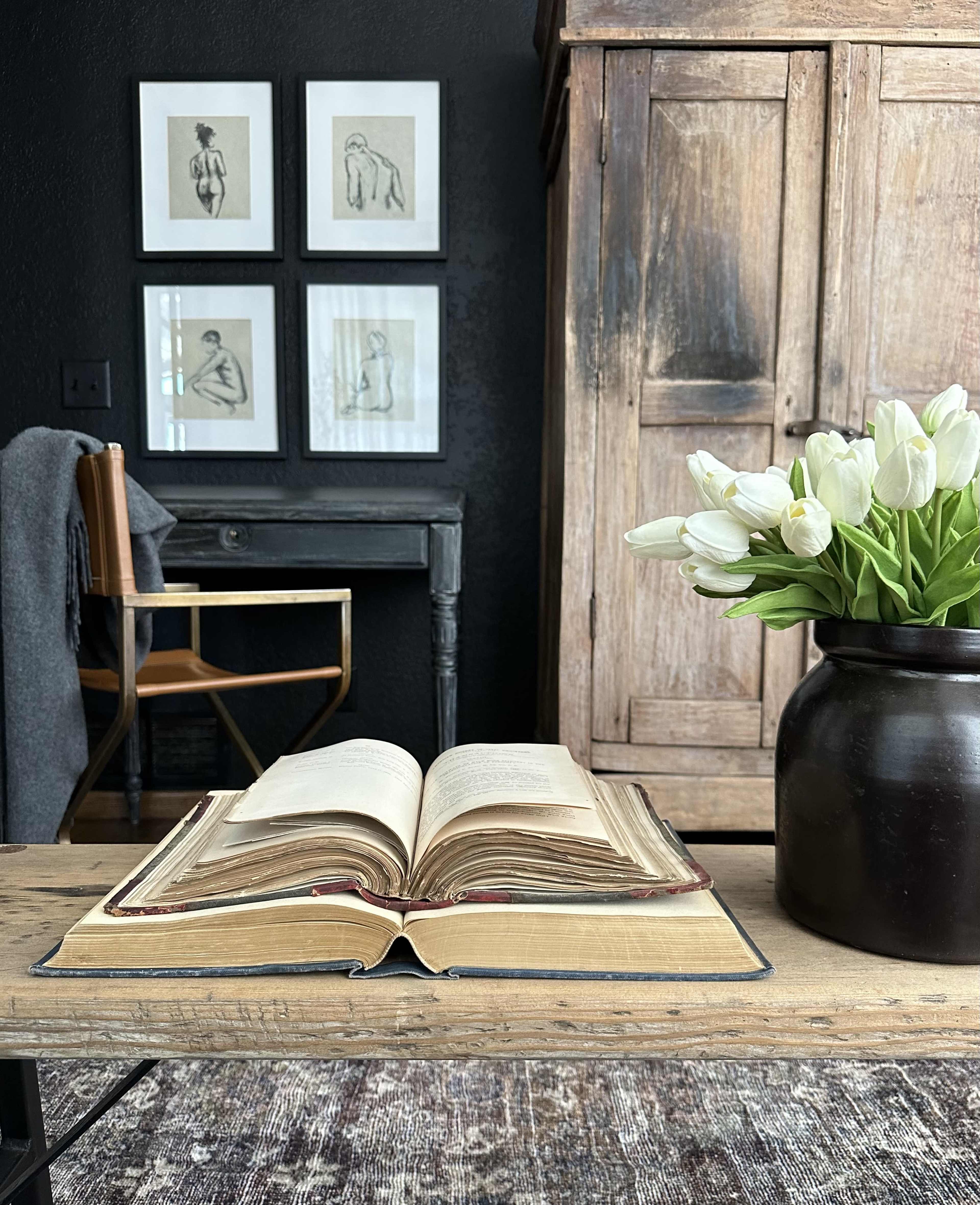An open book rests on a wooden table in a room featuring art on the wall, a chair, and a vase of white tulips.