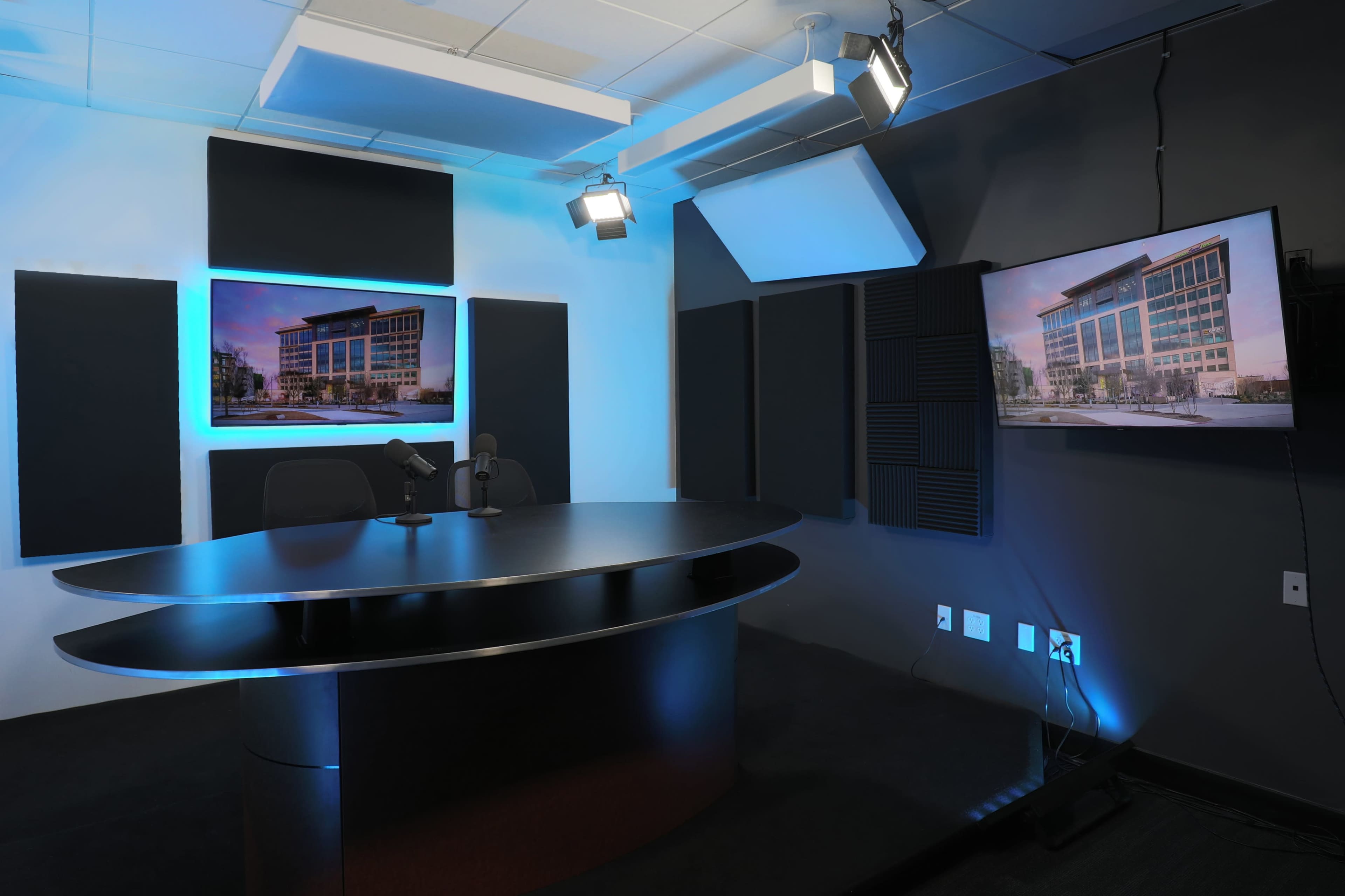 A modern recording studio featuring a round table, two microphones, soundproof panels, and a television displaying a building exterior.