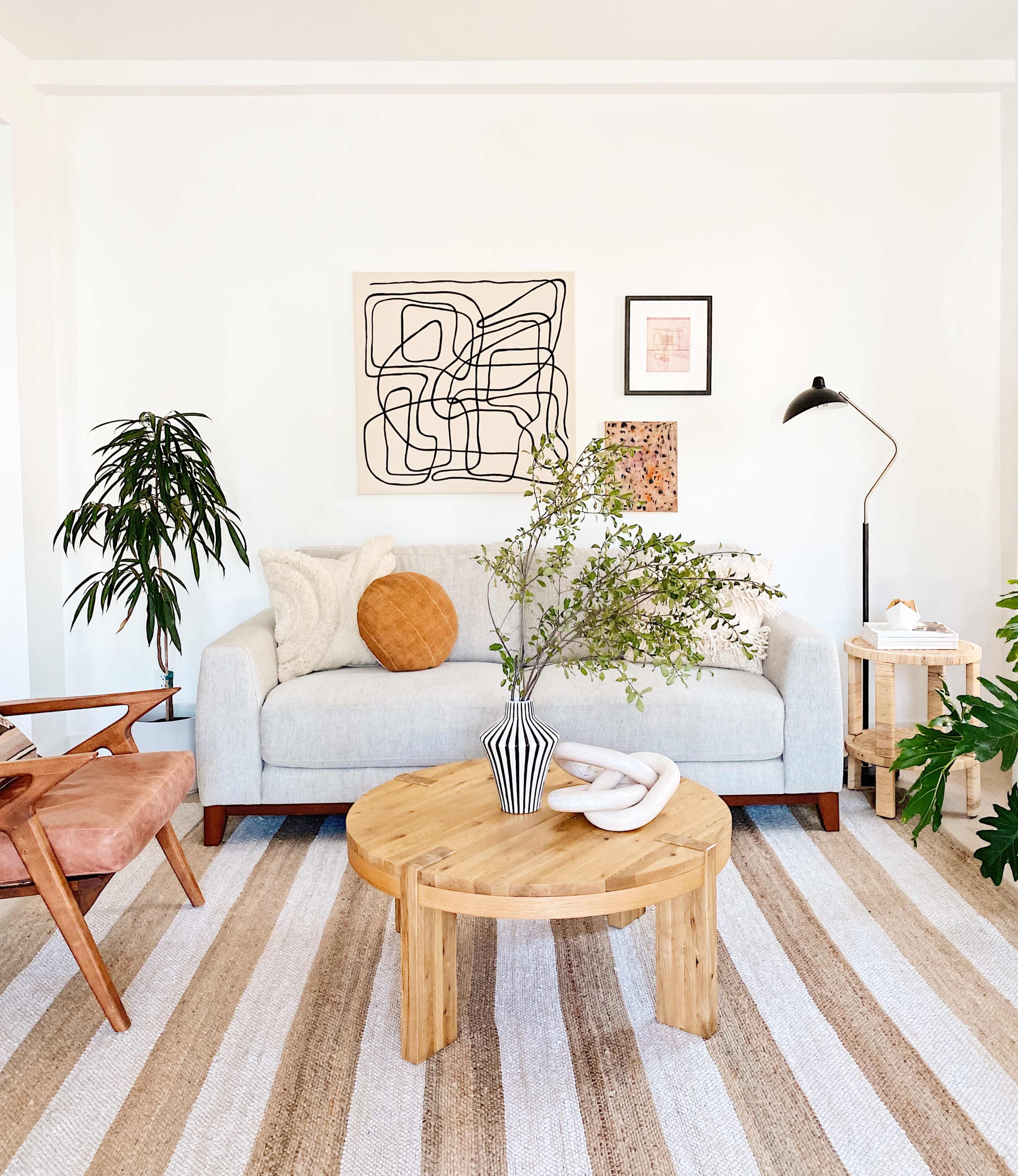 A modern living room features a light gray sofa, a round wooden coffee table, a potted plant, and an abstract artwork on the wall.